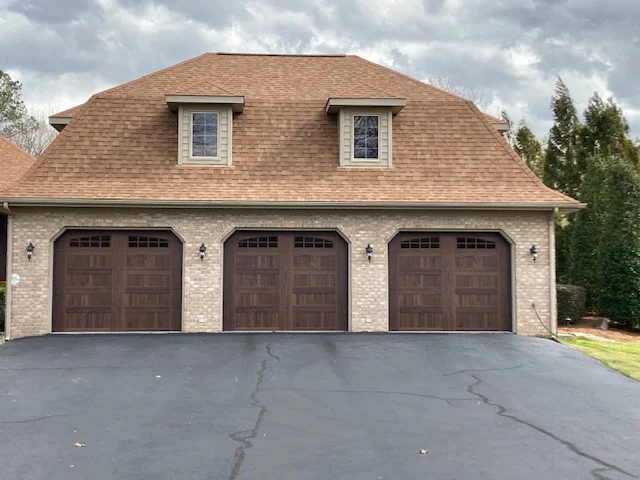 Three-car garage with tan shingled roof and dark brown doors, fronted by a wide driveway.