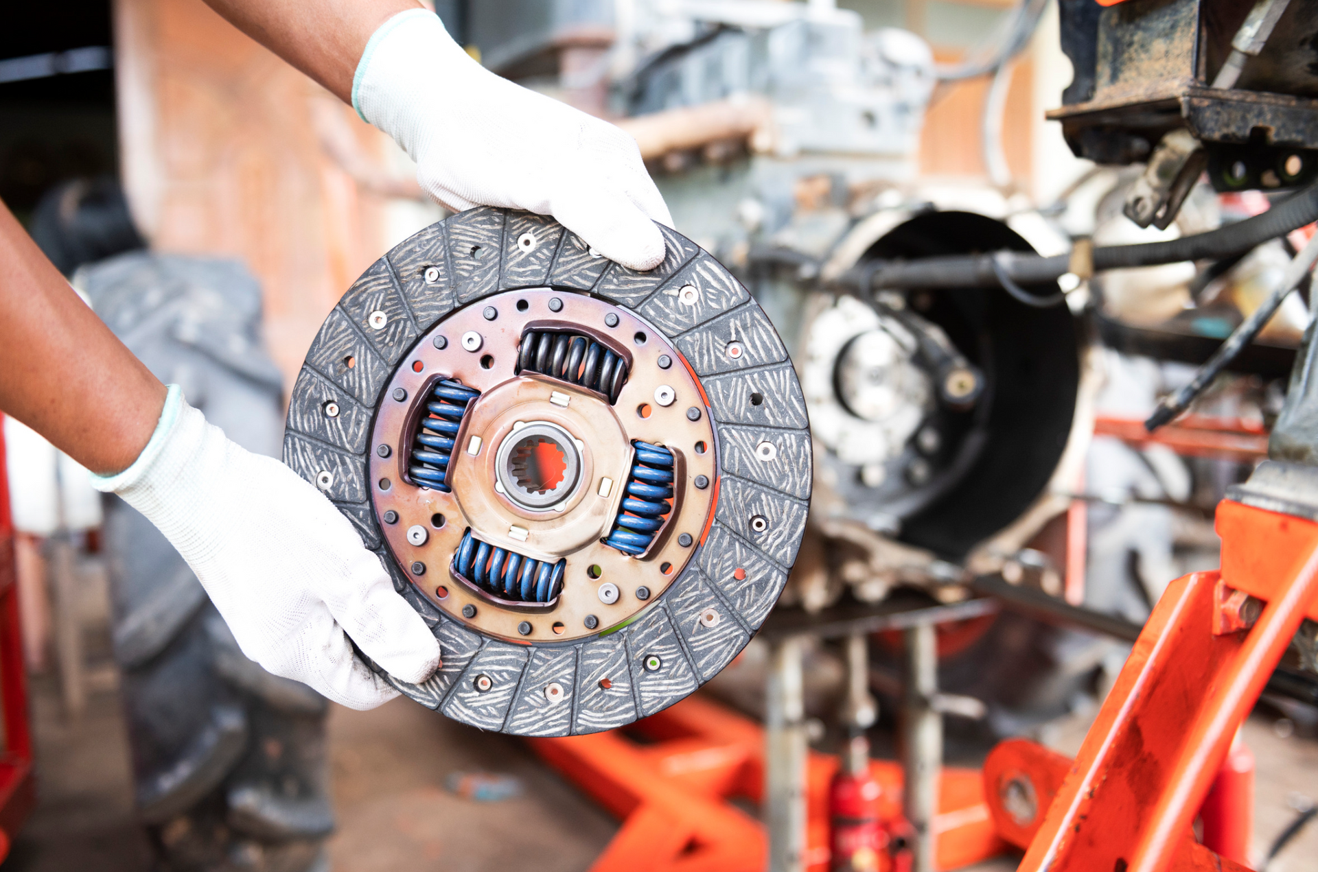 A person is holding a clutch disc in their hands in front of a car engine.