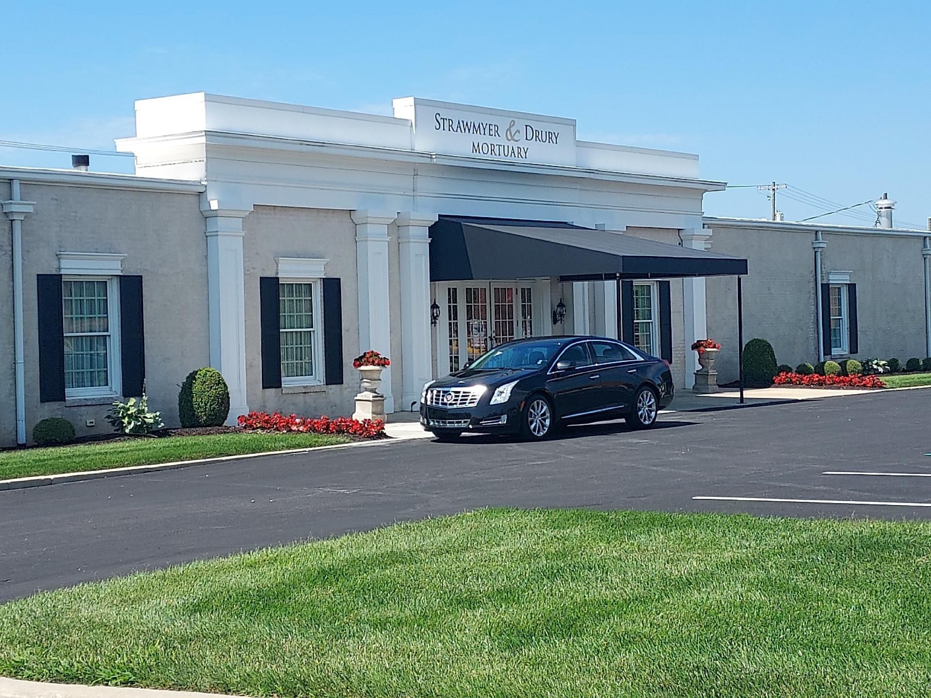 Front entrance of a white building with columns, a black car parked outside, and flower beds along the drive.