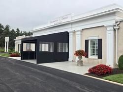 Building entrance with white columns, black canopy, and red flower planters along a paved driveway
