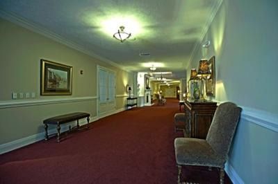 Long hotel hallway with red carpet, white walls, chandeliers, and chairs along the side.