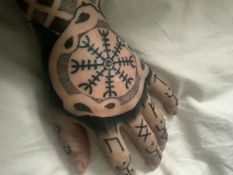 A hand with a large tattoo of the Helm of Awe symbol surrounded by a circular border, with runic symbols on the fingers.