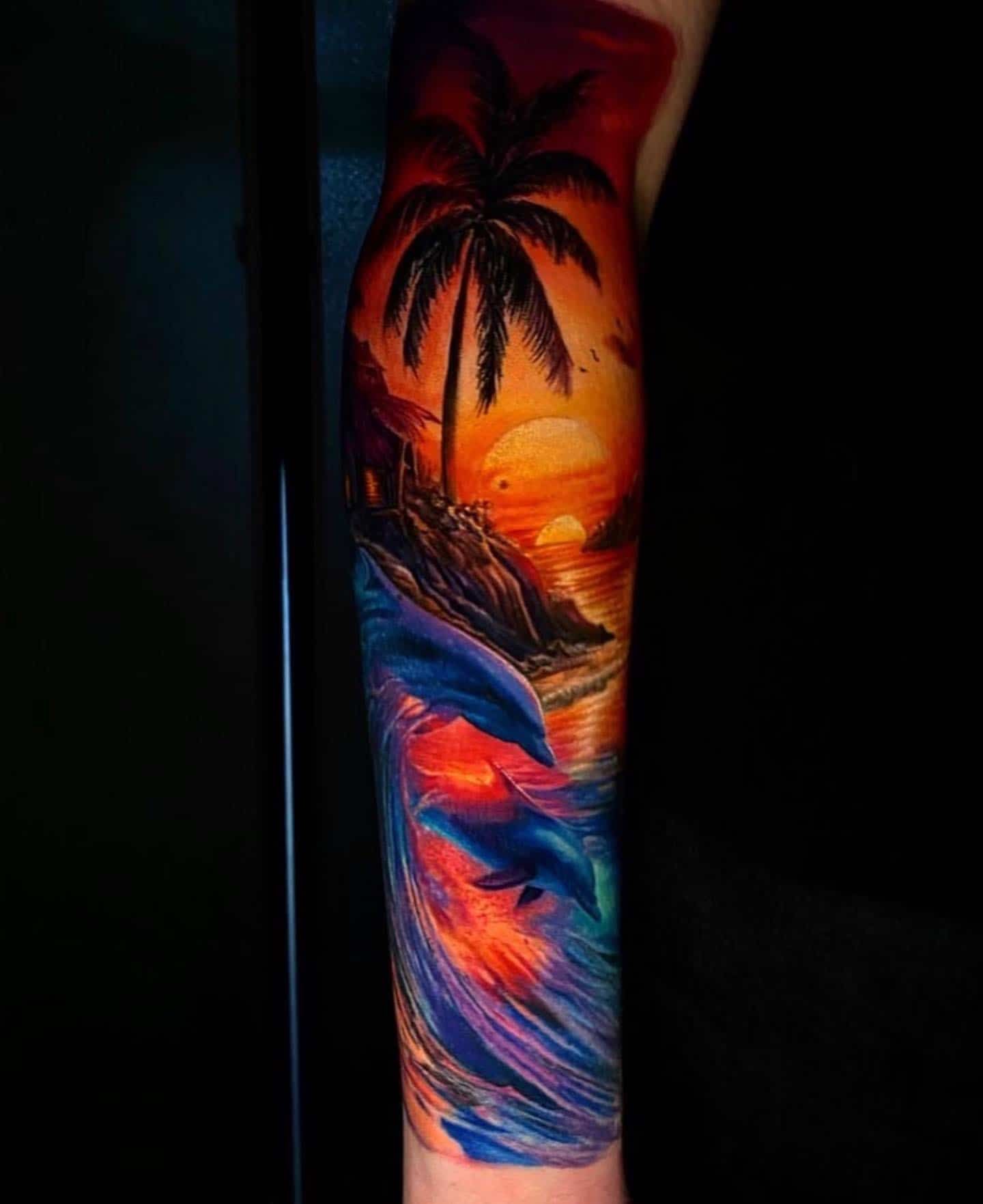 A vivid sunset tattoo on an arm, featuring a palm tree silhouette, a golden sun, and leaping blue dolphins in the waves.