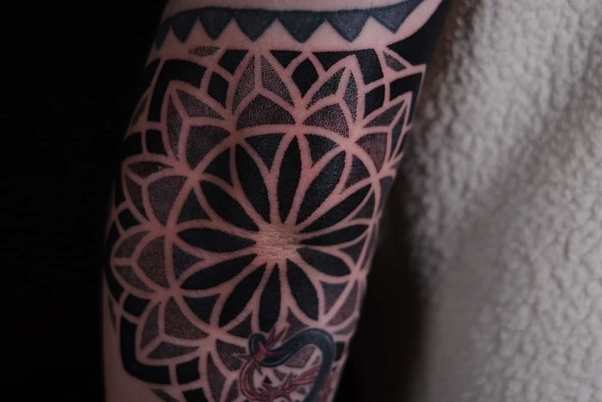 A close-up of a black ink geometric mandala tattoo on an elbow, featuring stippled shading and bold, symmetrical lines.
