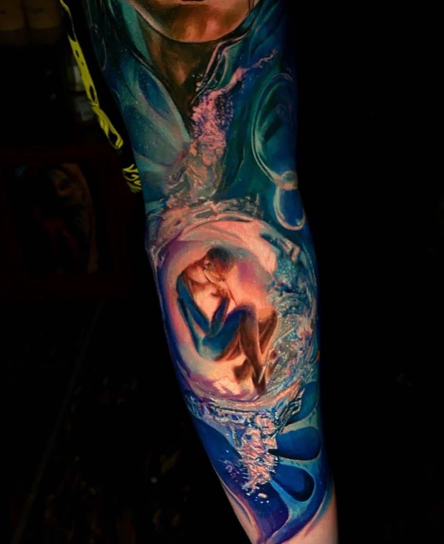 A vibrant tattoo of a figure curled in a glowing, iridescent bubble surrounded by flowing blue water and dark space.