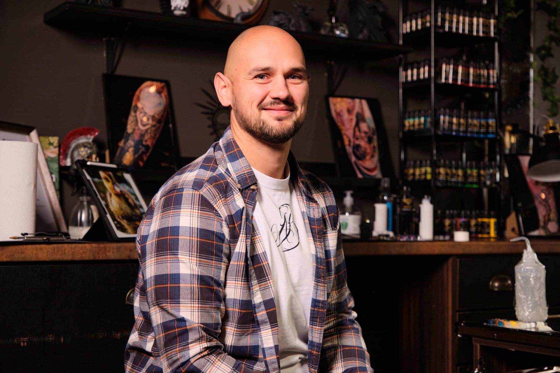 A person in a plaid shirt smiles in a tattoo studio filled with ink bottles and artwork displays.