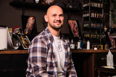 A smiling bald man in a plaid shirt sits in a tattoo studio filled with ink bottles and framed tattoo art.