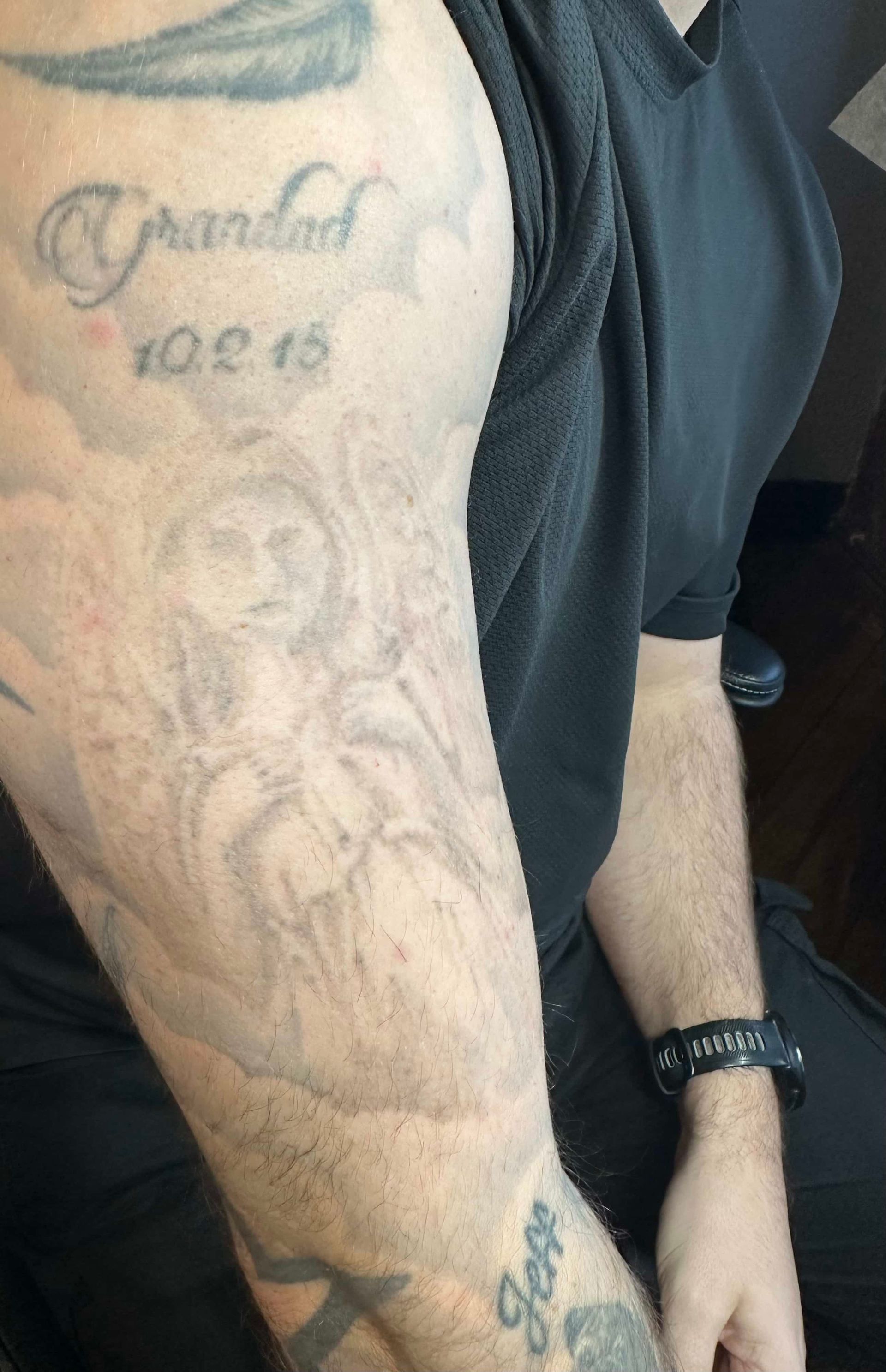 A person’s arm featuring multiple tattoos, including a portrait, clouds, text, and a date.