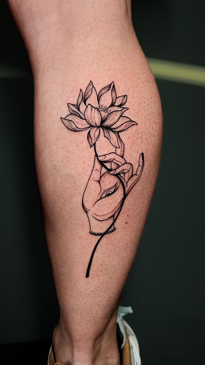 A black-line tattoo on a lower leg featuring a hand delicately holding a blooming lotus flower.