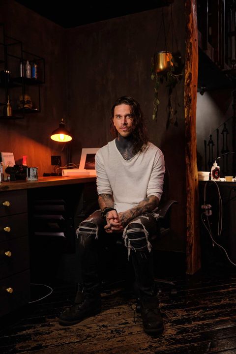 A person with neck tattoos, wearing a white long-sleeve shirt and distressed black jeans, sits in a dark, rustic studio.