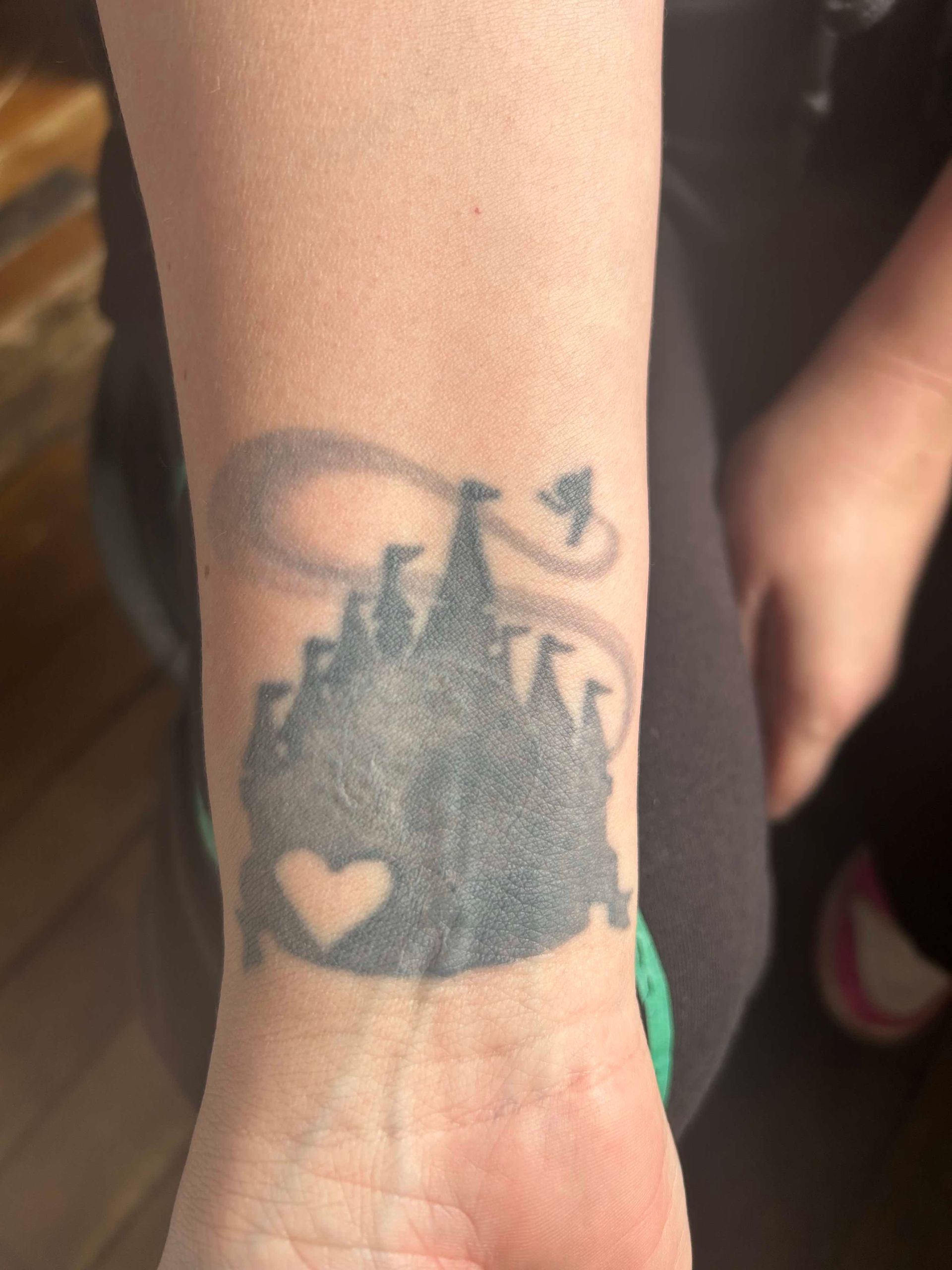 A wrist tattoo of a black silhouette of a Disney-style castle with a small white heart and Tinker Bell flying above.