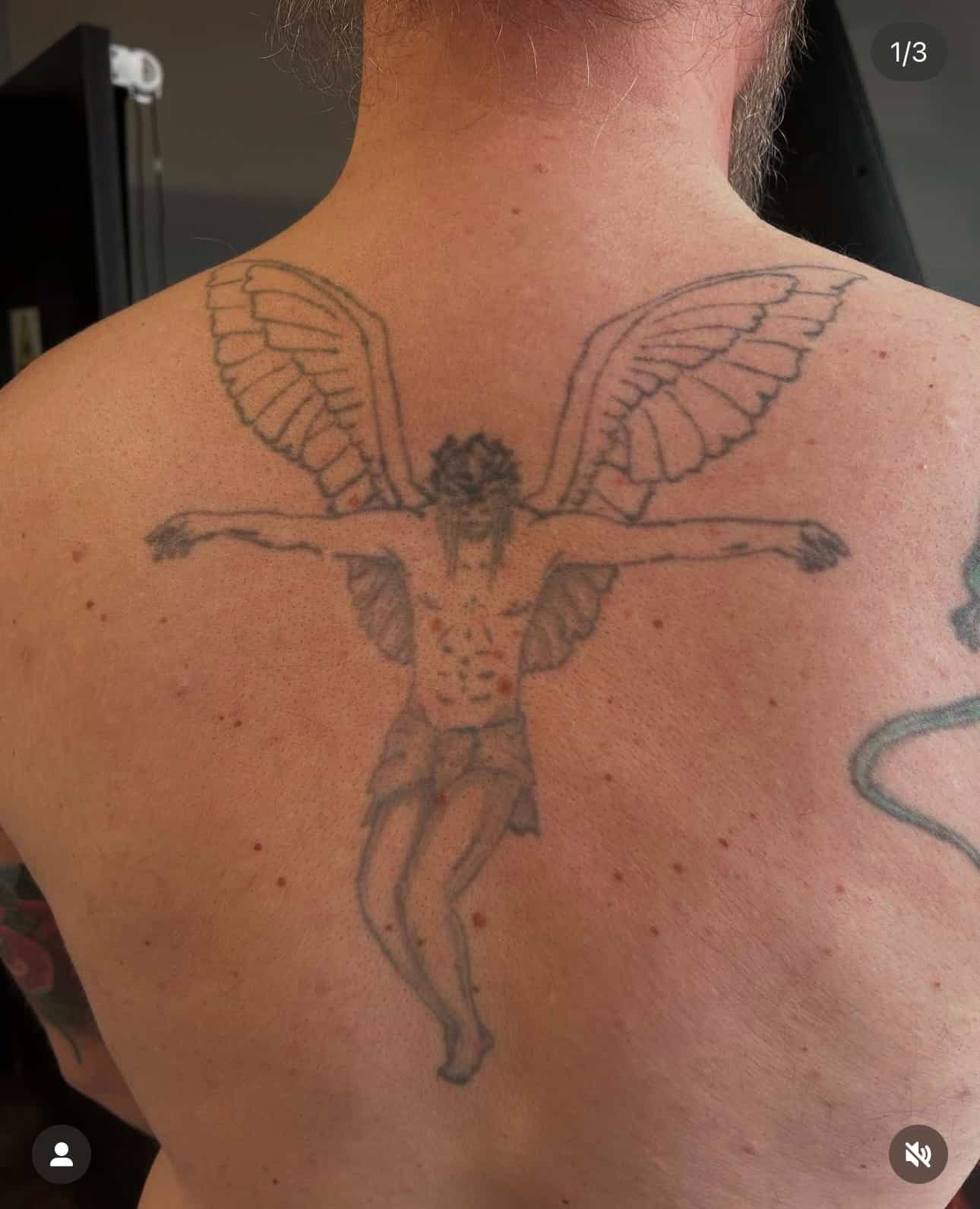A line-art tattoo on a person's upper back depicts a winged, crucified figure wearing a crown of thorns.