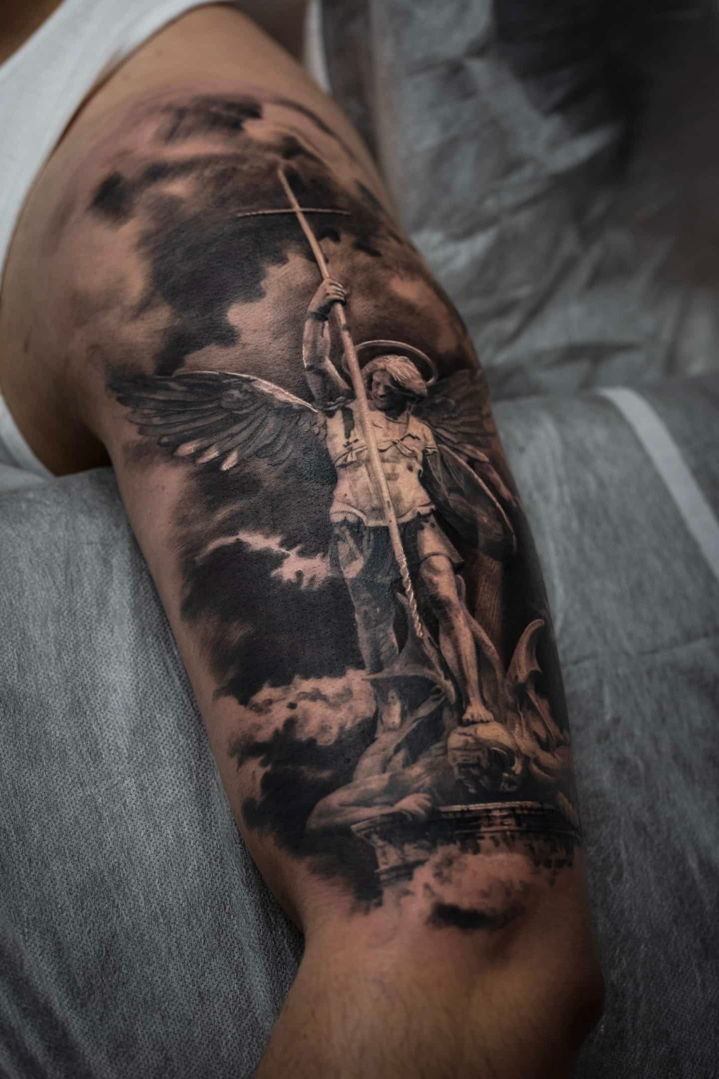 A black-and-grey tattoo on an upper arm depicting an angel with wings wielding a spear amidst dark, stormy clouds.