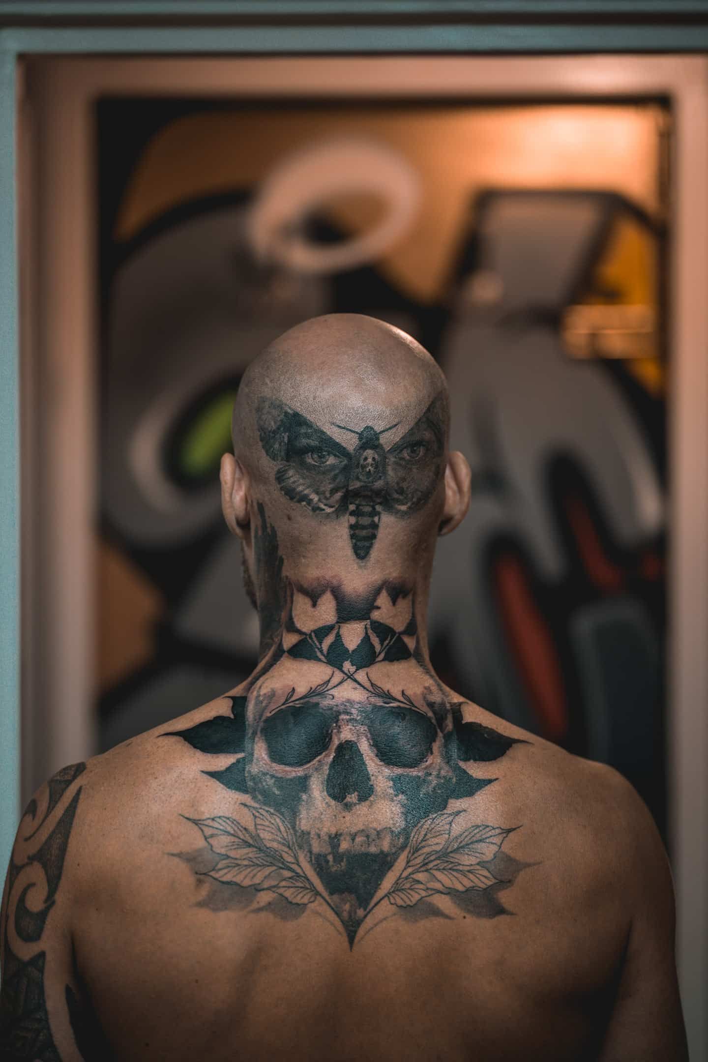 A person seen from behind, featuring an intricate moth tattoo on their scalp and a large skull tattoo on their upper back.