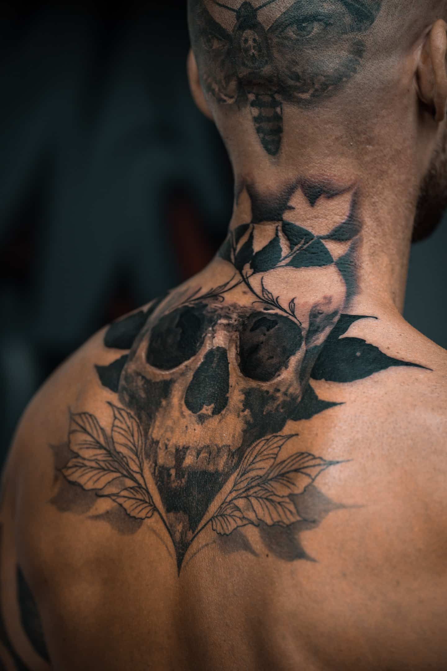 A close-up of a person's upper back and neck featuring a large, dark, detailed skull tattoo surrounded by leaf designs.