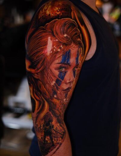 A realistic upper-arm tattoo depicting a person with blue face paint, long hair, and a small silhouette of a warrior below.