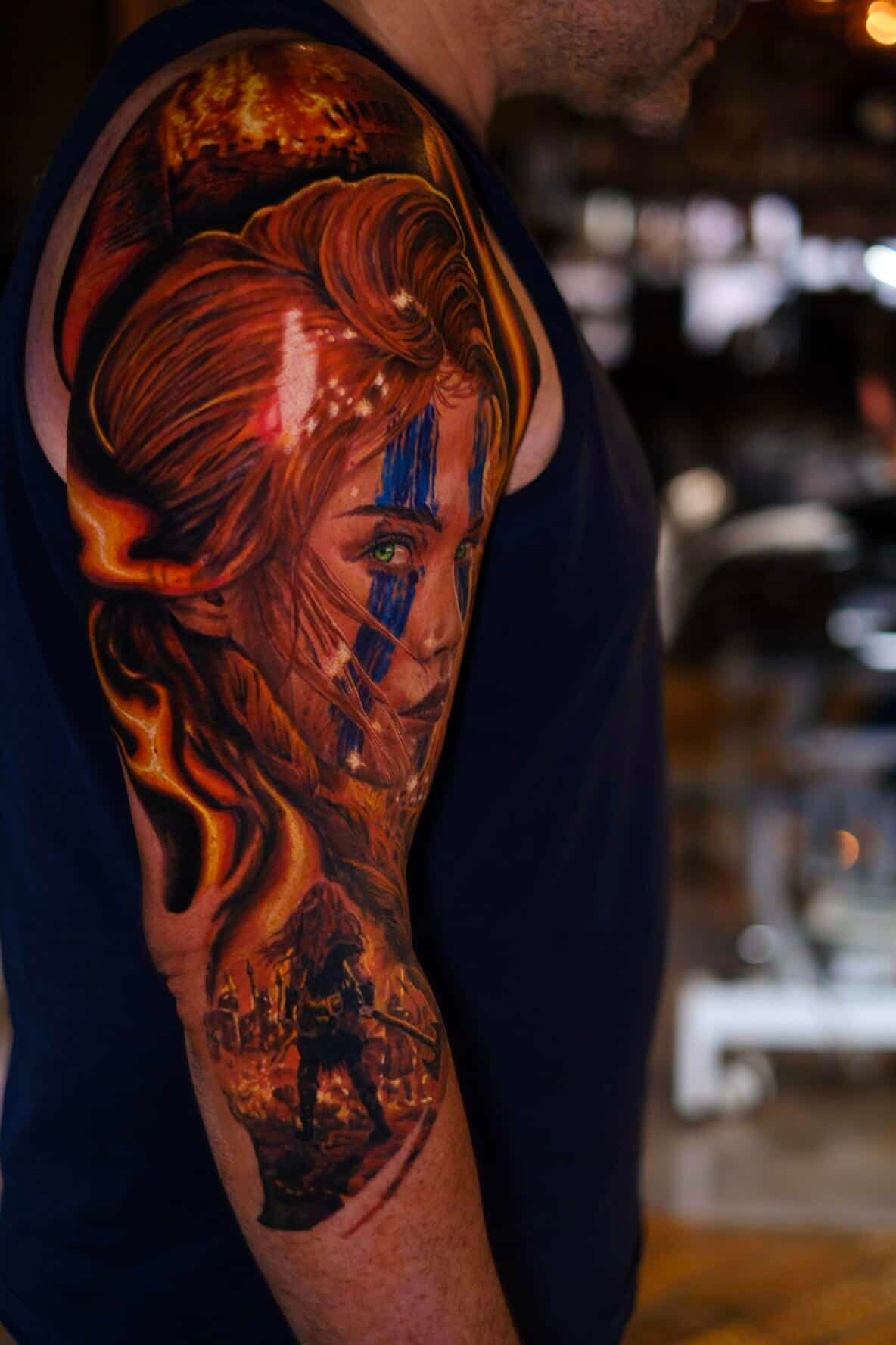 A vibrant, flame-themed tattoo on an upper arm featuring a warrior-like figure with blue facial markings.