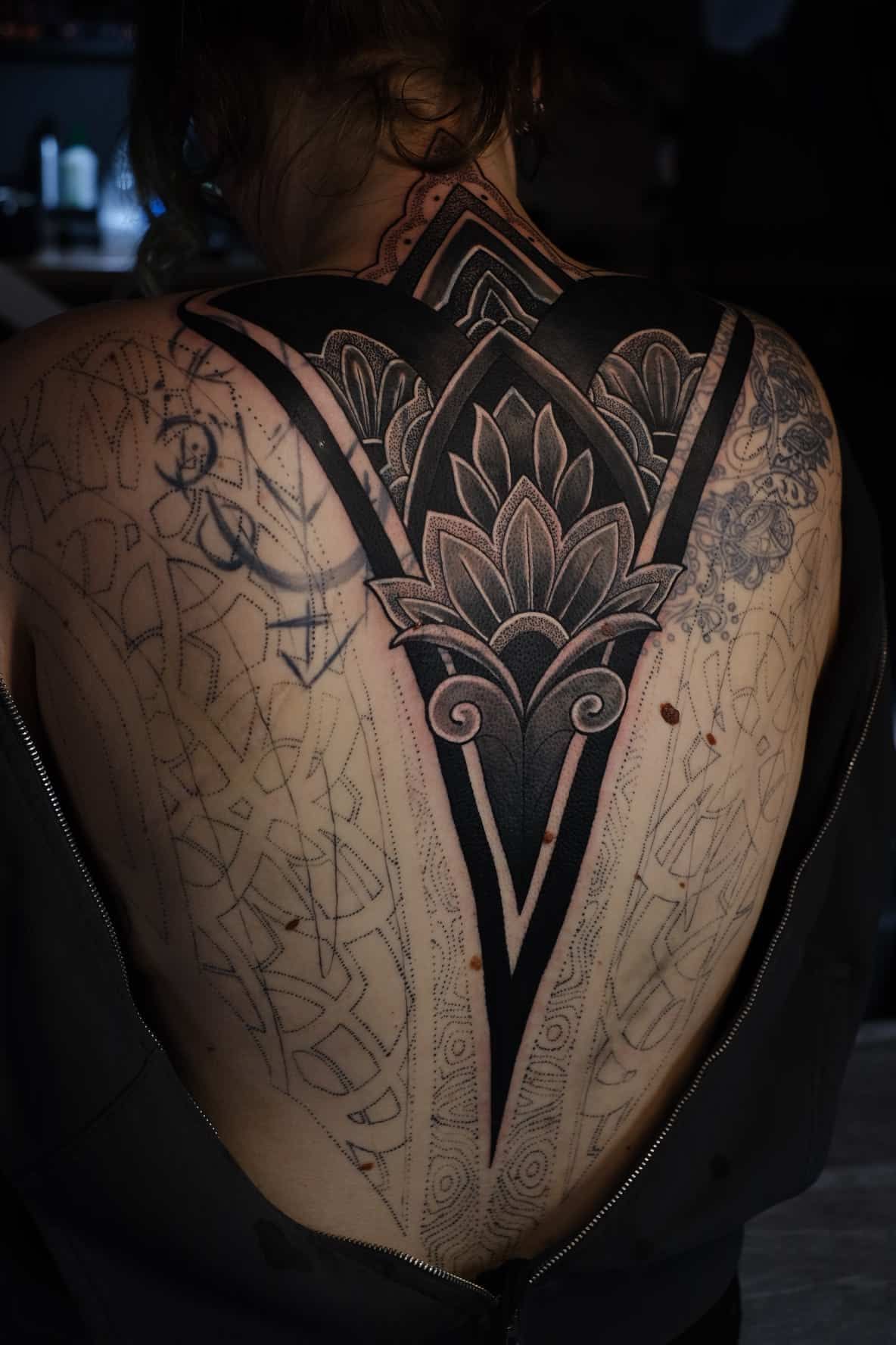 A person’s back with a large, intricate black-ink tattoo featuring floral and geometric patterns centered on the spine.