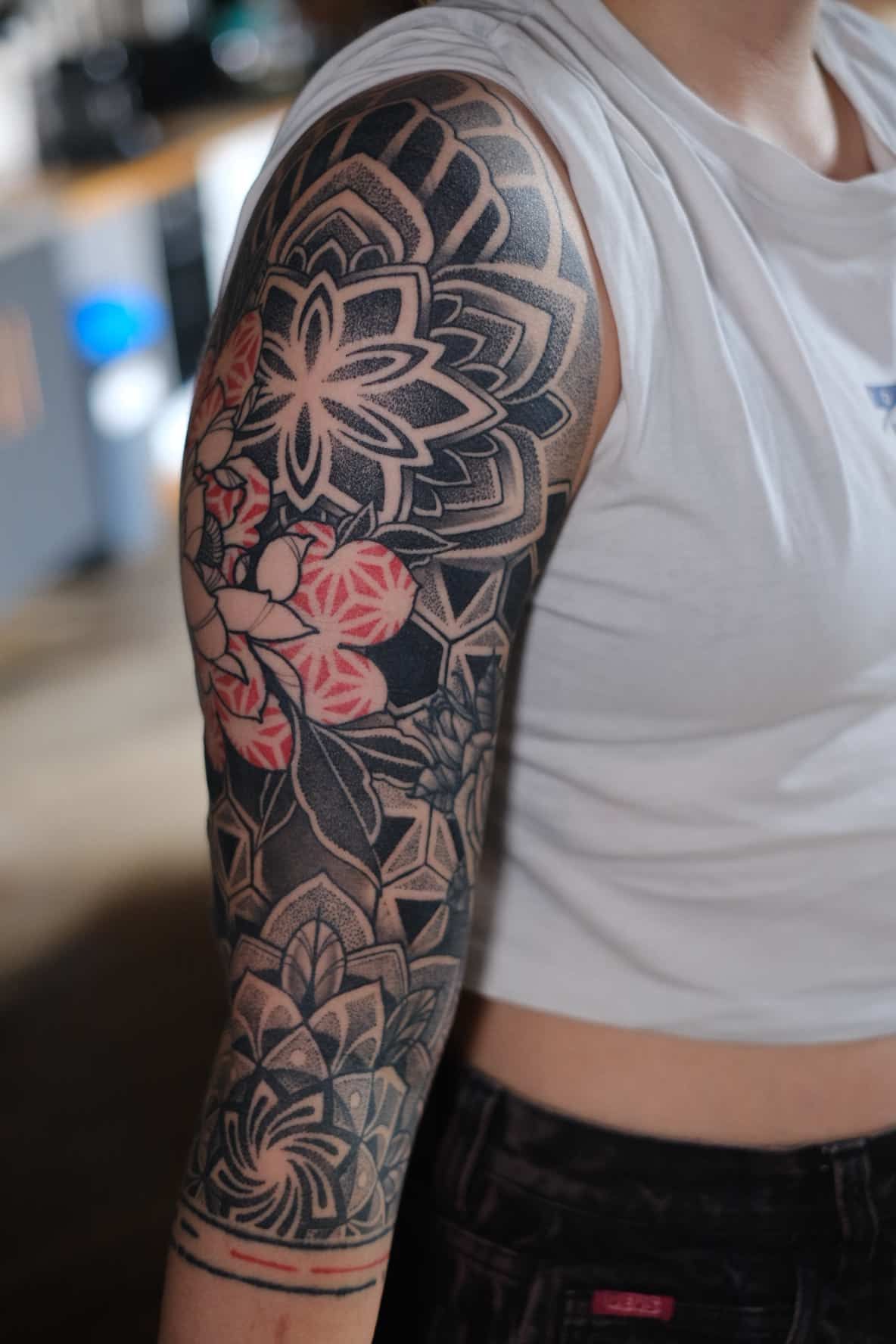 A person’s arm featuring an intricate black-ink mandala and geometric sleeve tattoo with red floral accents.