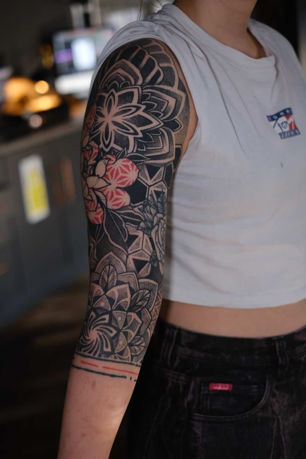A person wears a dark, intricate arm sleeve tattoo featuring repeating mandala patterns and small red flower accents.