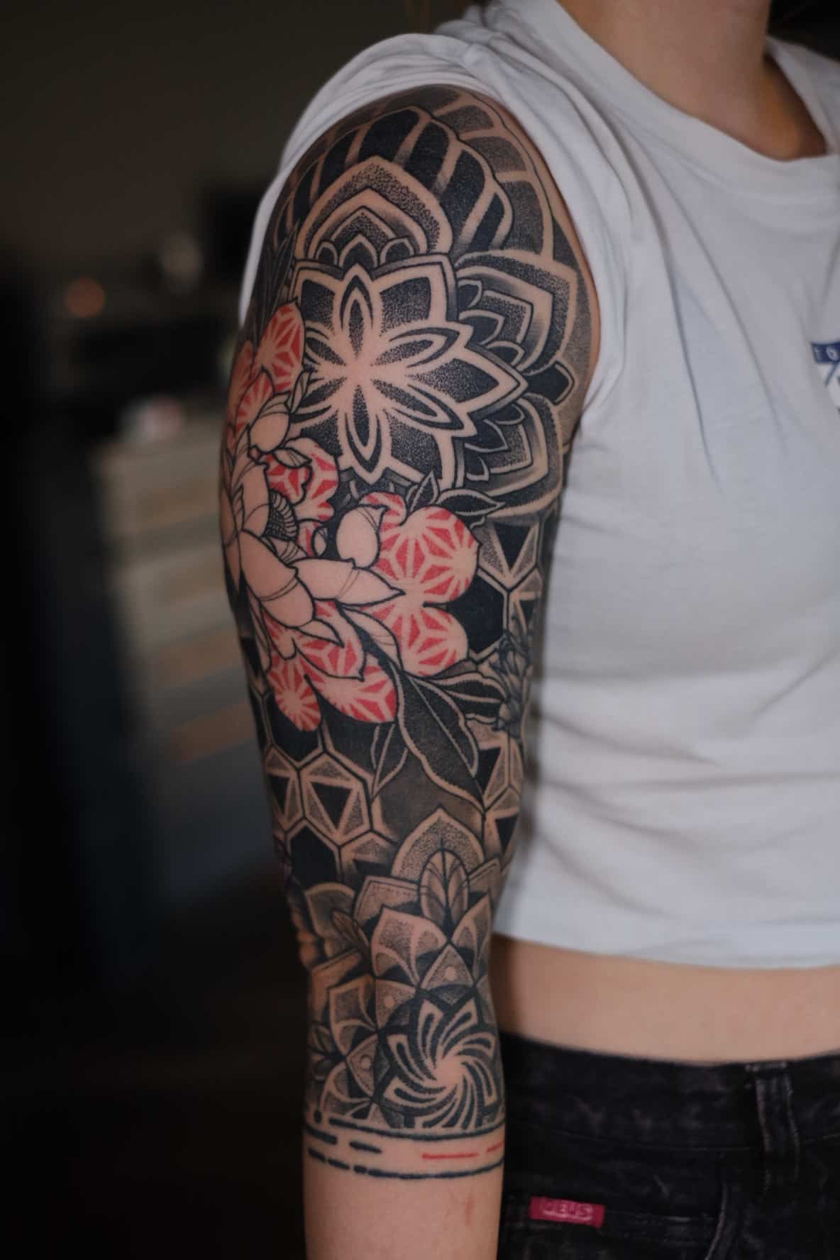 A person's upper arm featuring a blackwork tattoo with geometric patterns, mandalas, and red floral accents.