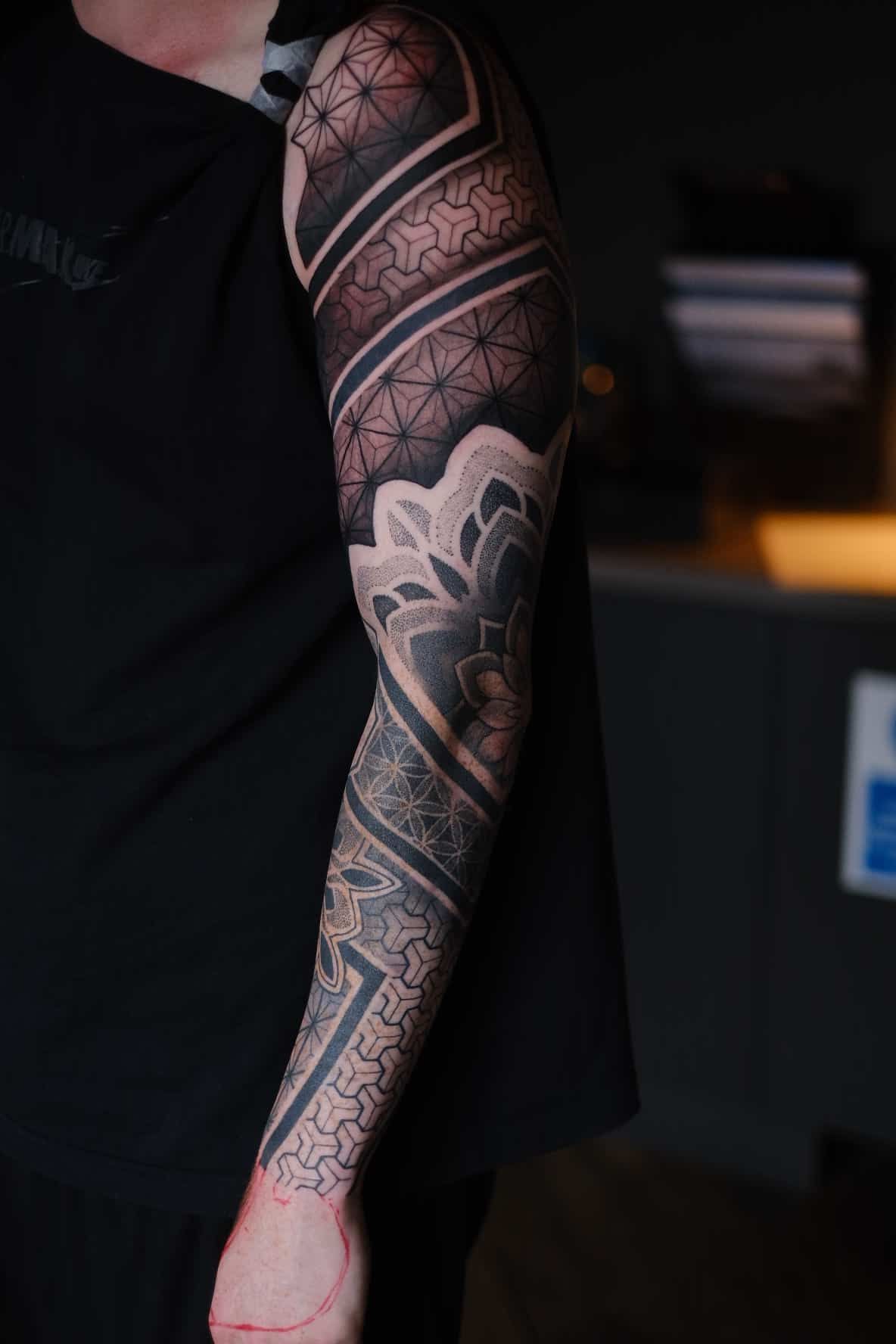 Arm sleeve tattoo featuring intricate geometric patterns, a central mandala design, and black-and-grey shading.