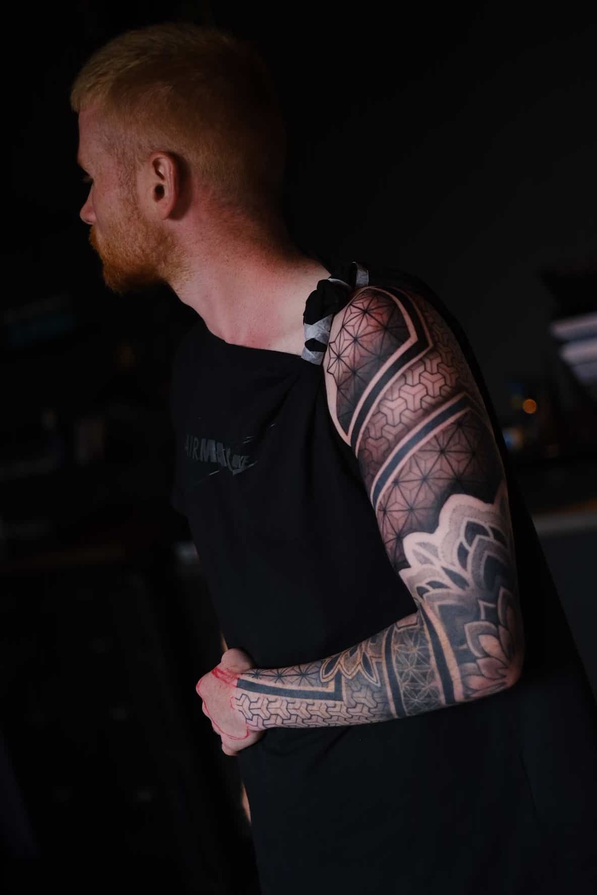 A person with a red beard wearing a black tank top, displaying a detailed black-and-grey geometric sleeve tattoo.