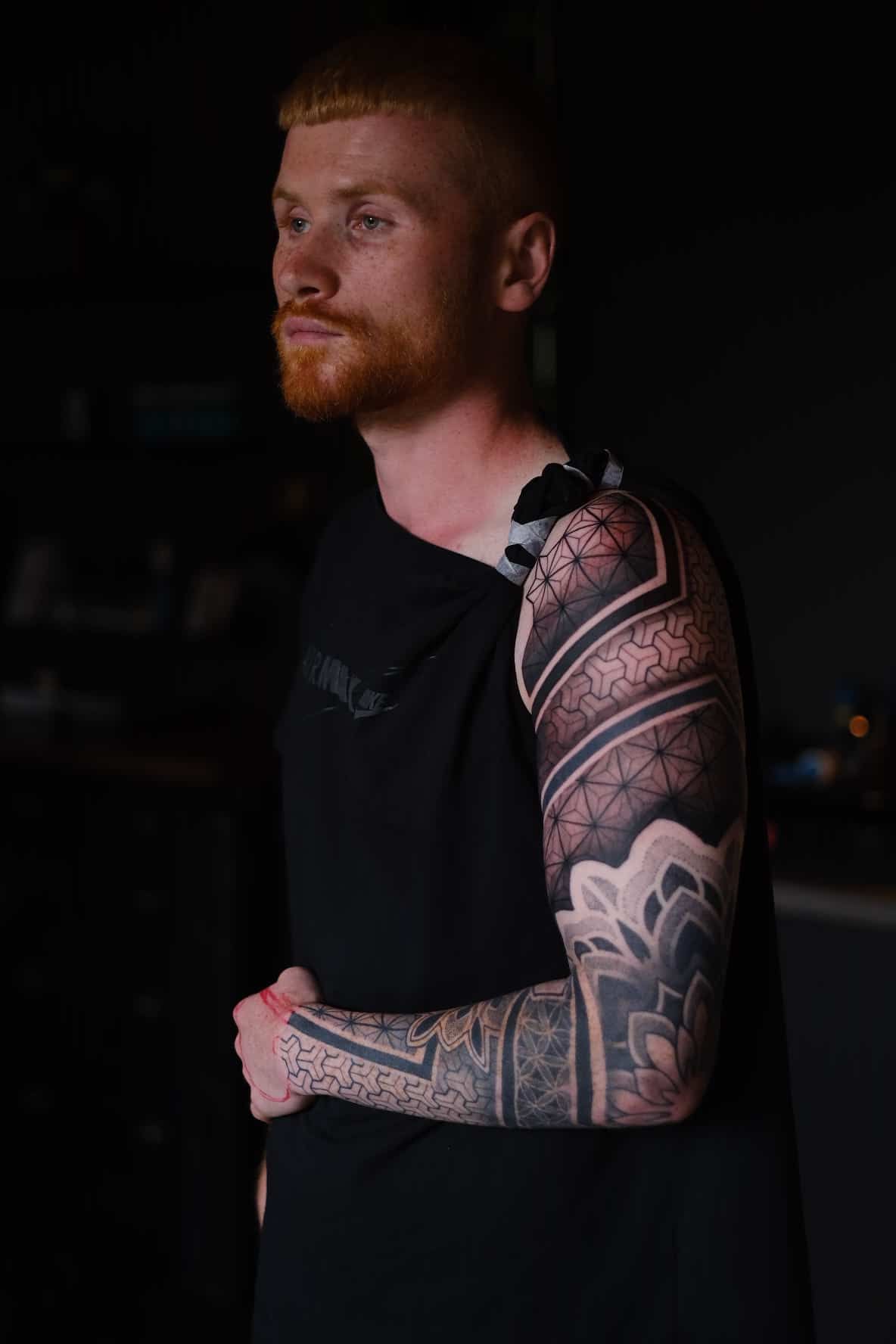 A person with red hair and a beard, wearing a black top, displays an intricate, geometric patterned sleeve tattoo.