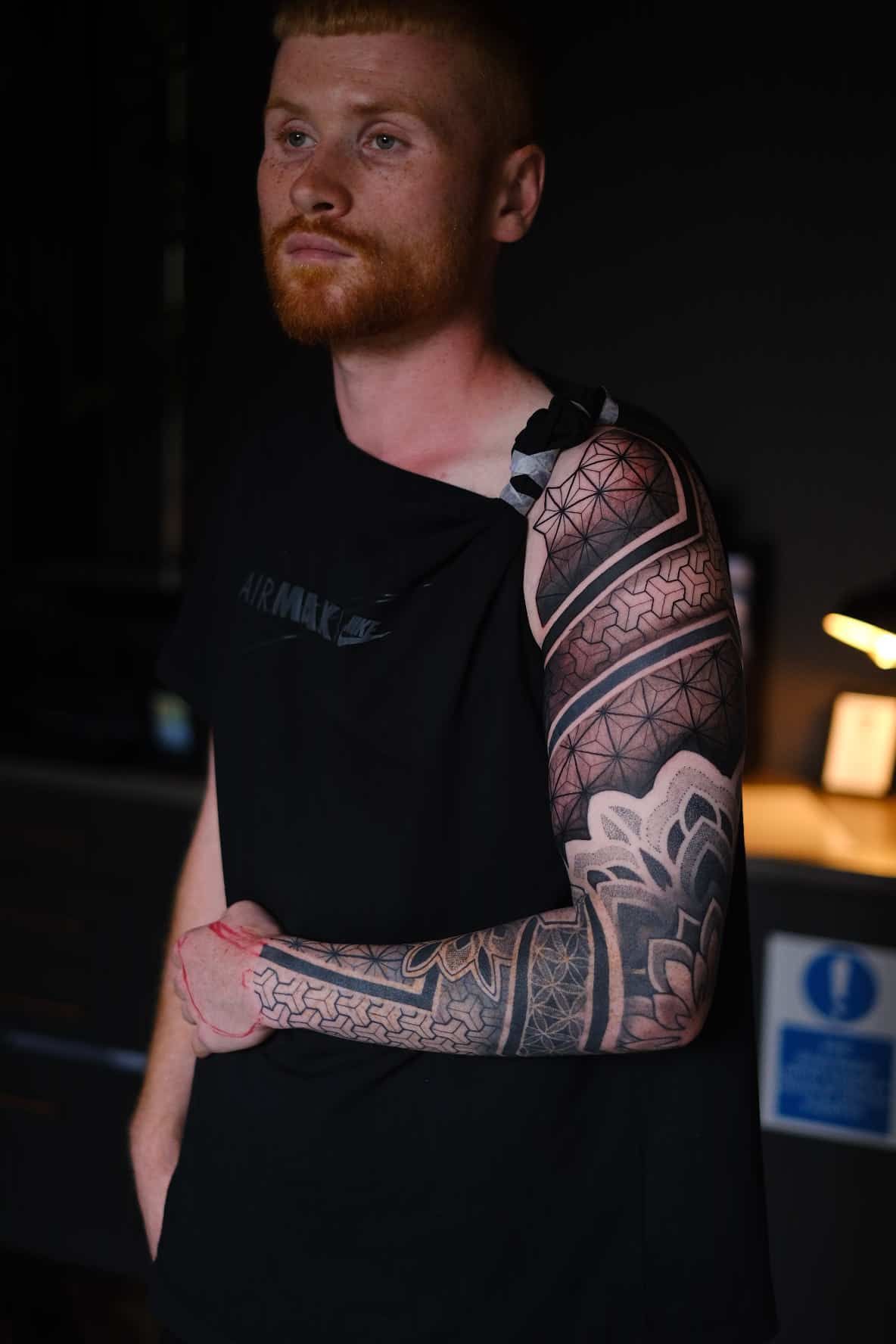 A person with a detailed geometric and mandala-style full sleeve tattoo stands against a dark background.