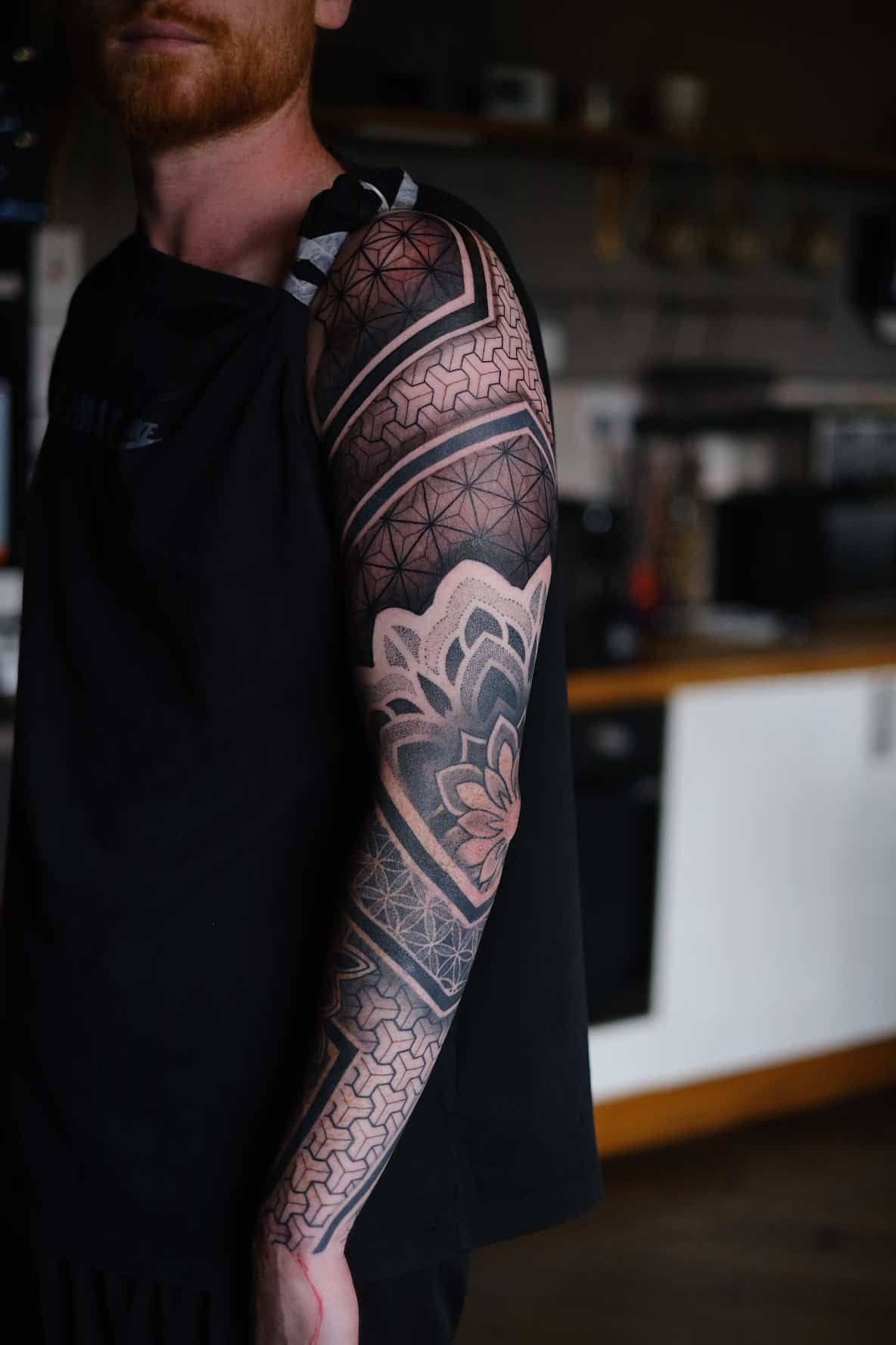 A person wearing a sleeveless black shirt, displaying an intricate geometric and mandala-patterned sleeve tattoo.