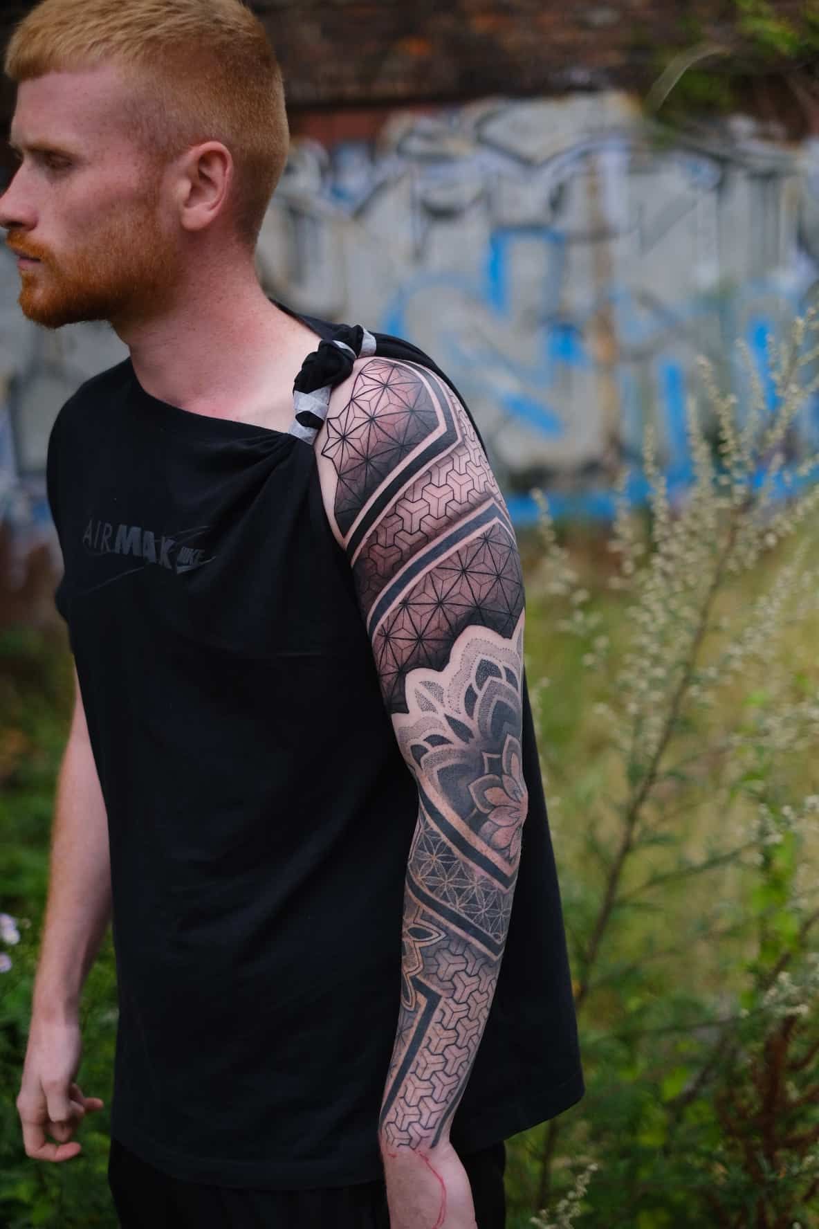 A person with ginger hair stands outdoors wearing a black tank top, displaying a detailed geometric tattoo on their arm.