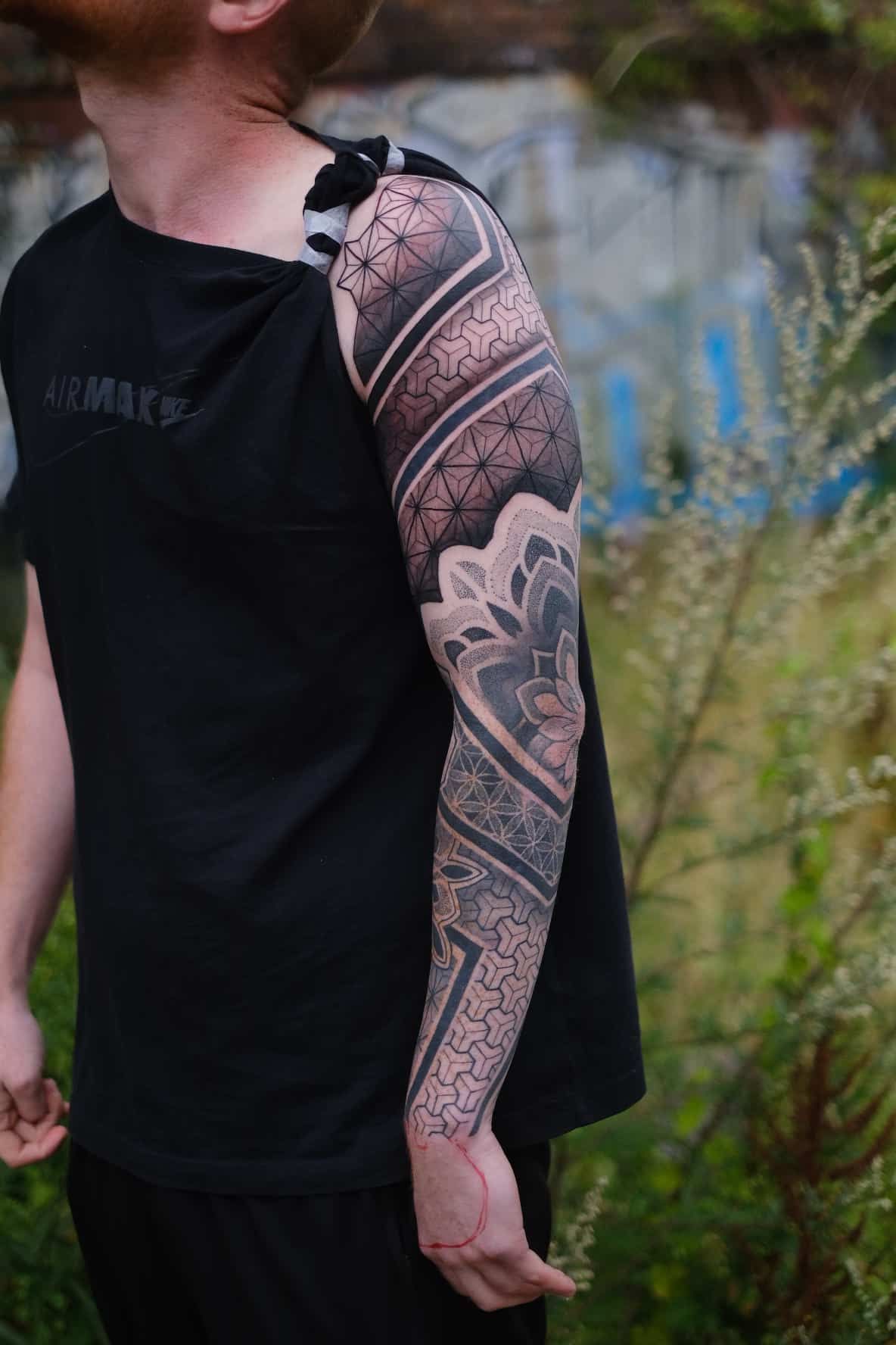 A person wearing a black shirt shows off a detailed blackwork sleeve tattoo with geometric and floral patterns.