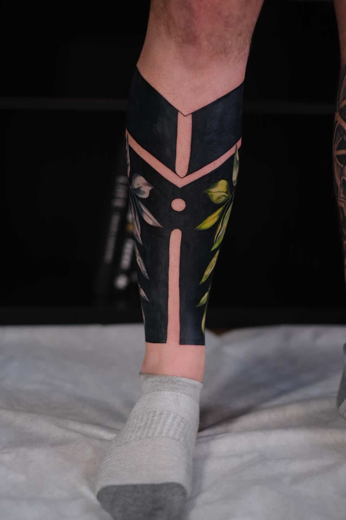 A lower leg featuring a blackwork tattoo with geometric cutouts and detailed botanical imagery.