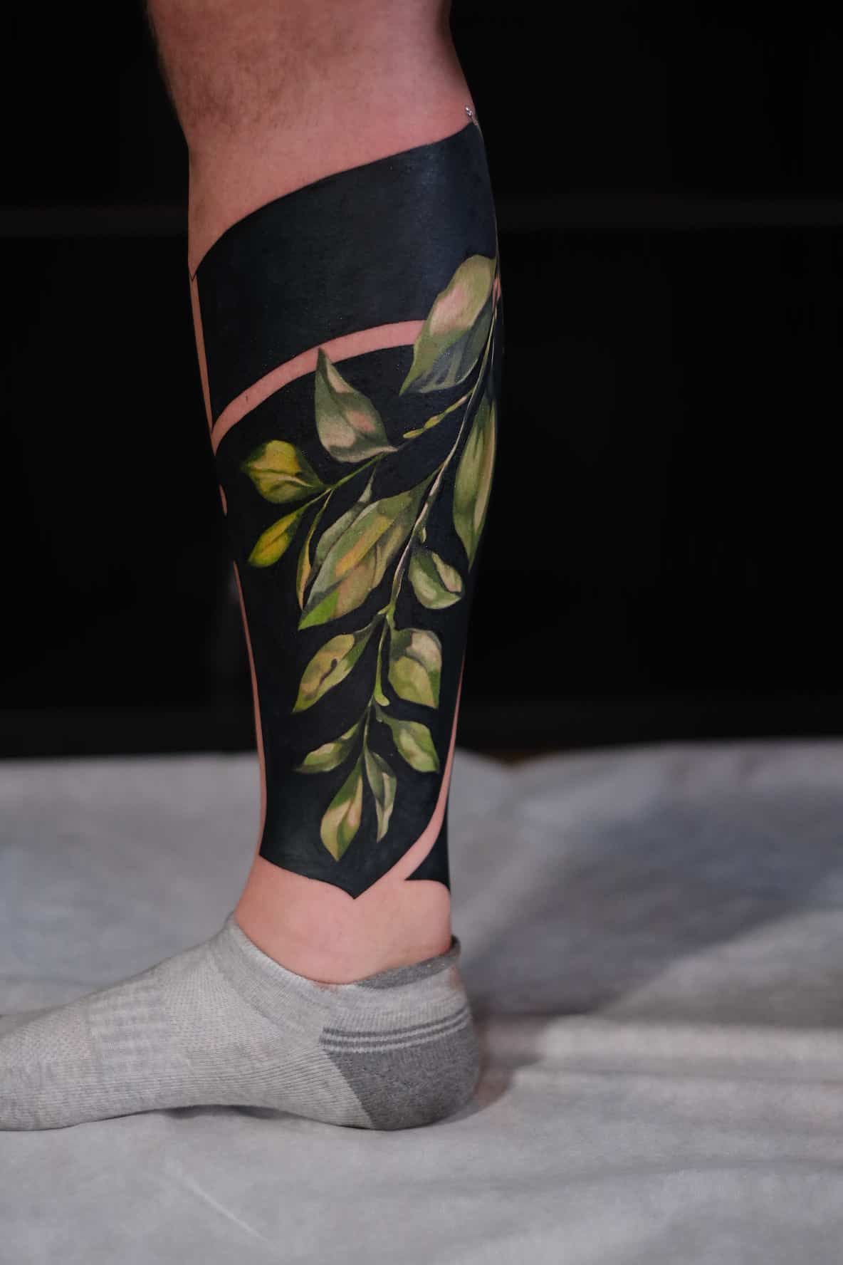 A lower leg tattoo featuring a solid black block design with a realistic botanical branch in shades of green.