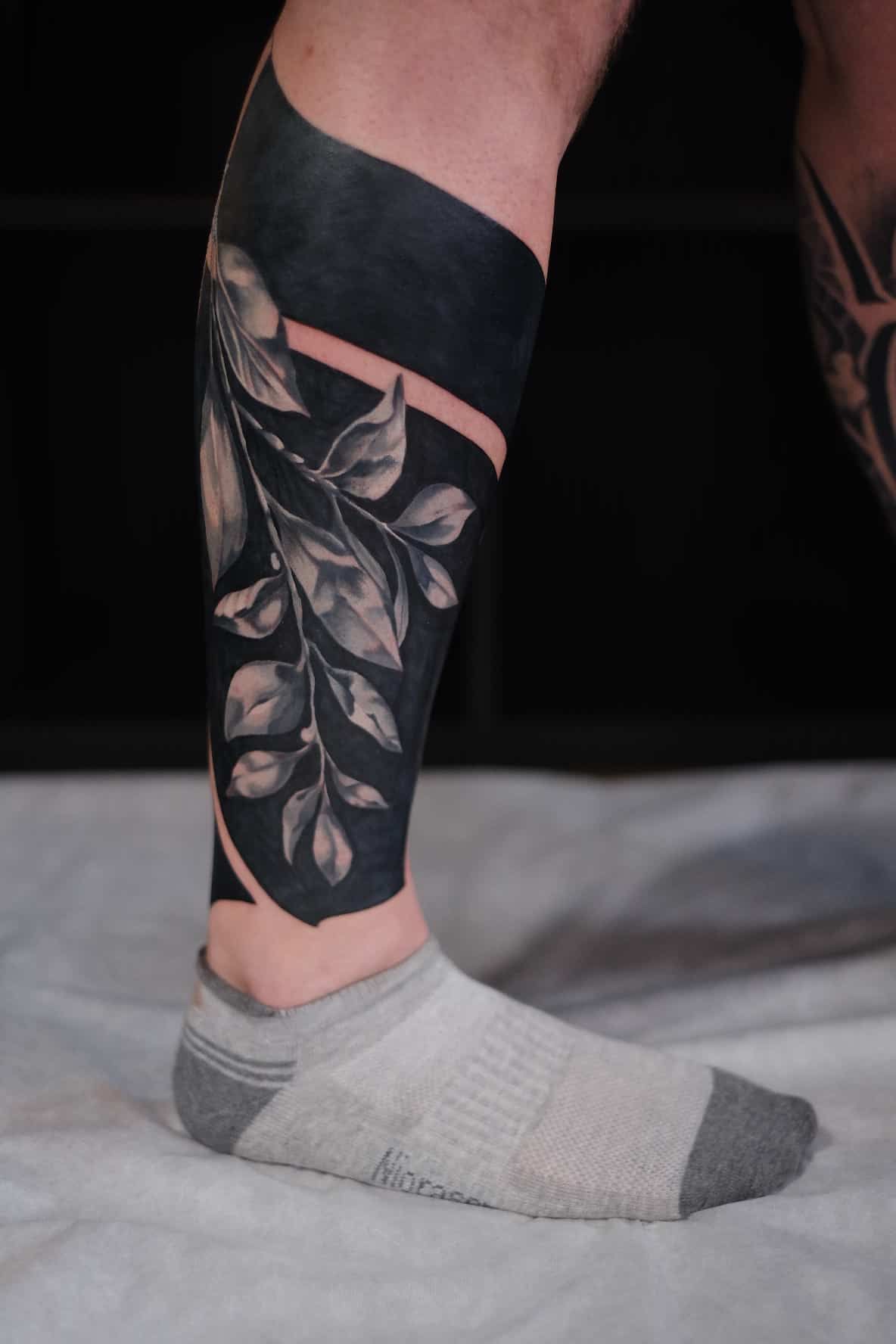 A lower leg tattoo featuring a solid black band with a negative-space cutout of delicate botanical leaves.