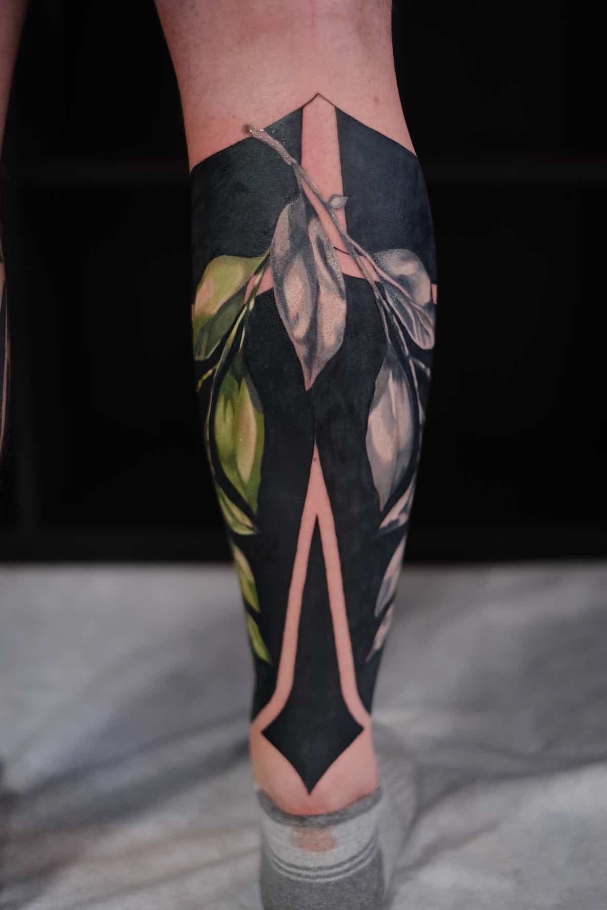 A lower leg tattoo featuring a solid black geometric design layered with detailed, light-colored leaves and branches.