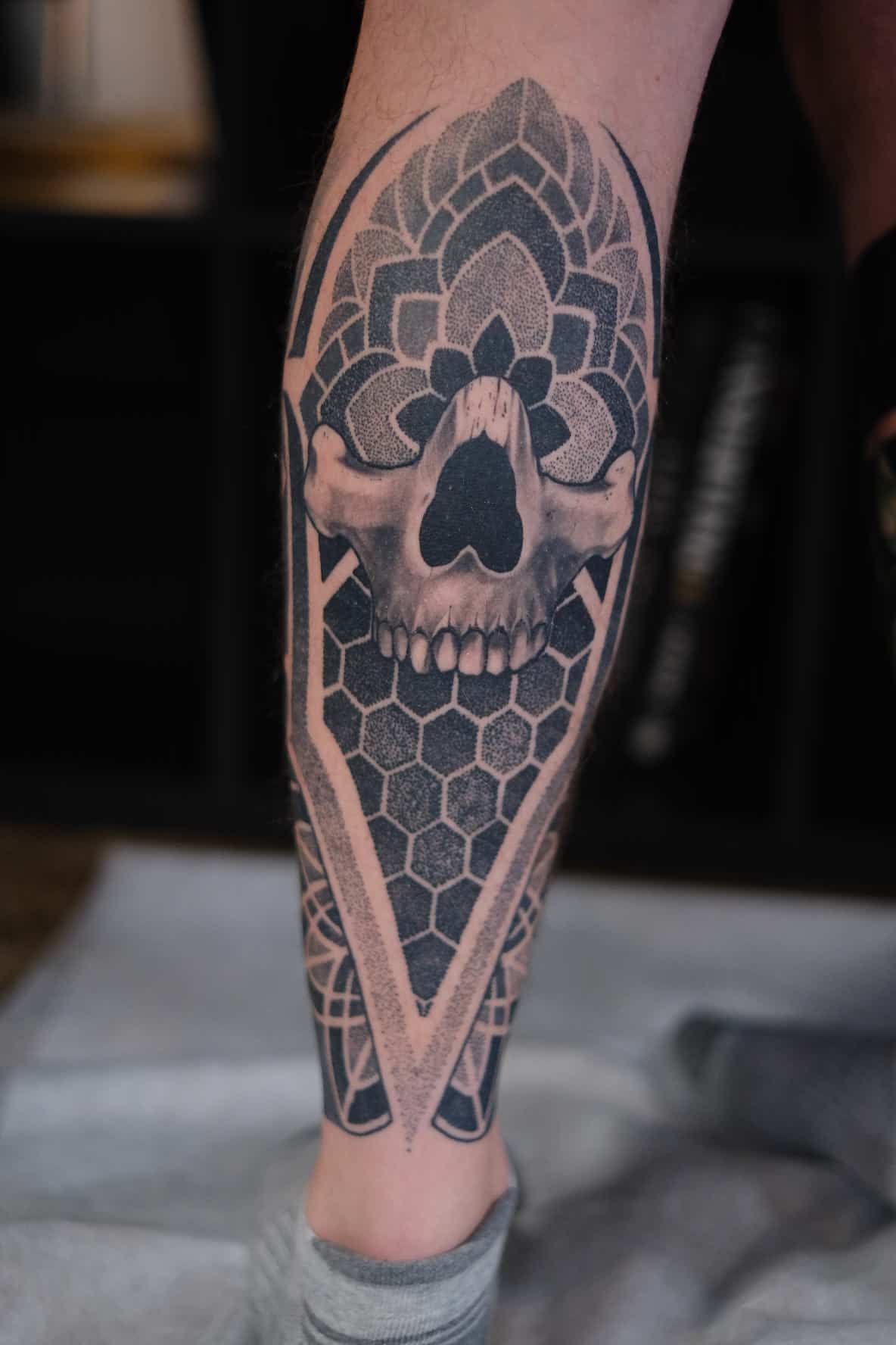 A tattoo on a lower leg featuring a central skull framed by geometric patterns, mandalas, and honeycomb shapes.