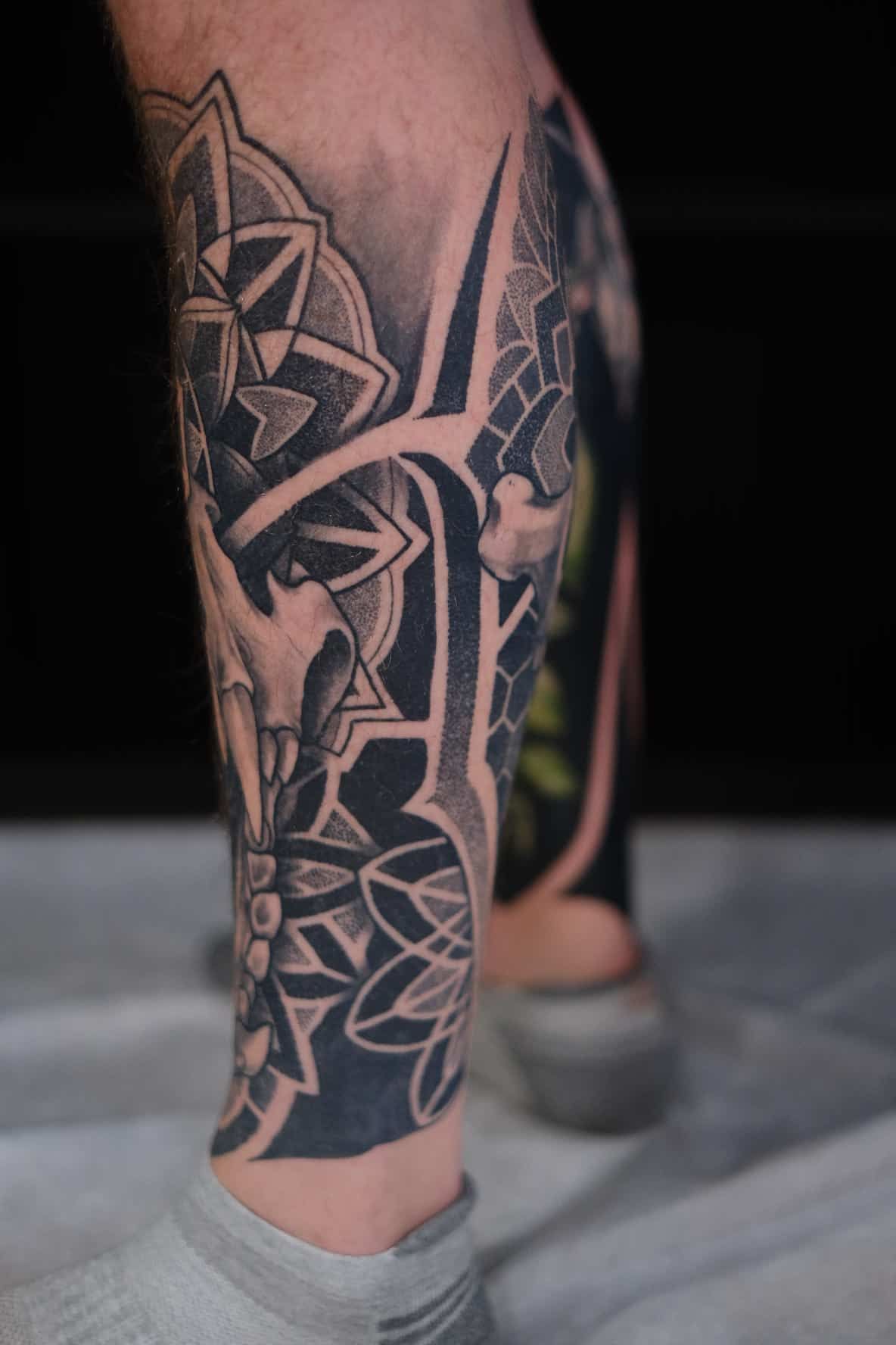 A black-and-grey tattoo on a lower leg featuring geometric mandala patterns, a skull motif, and bold, sharp linework.