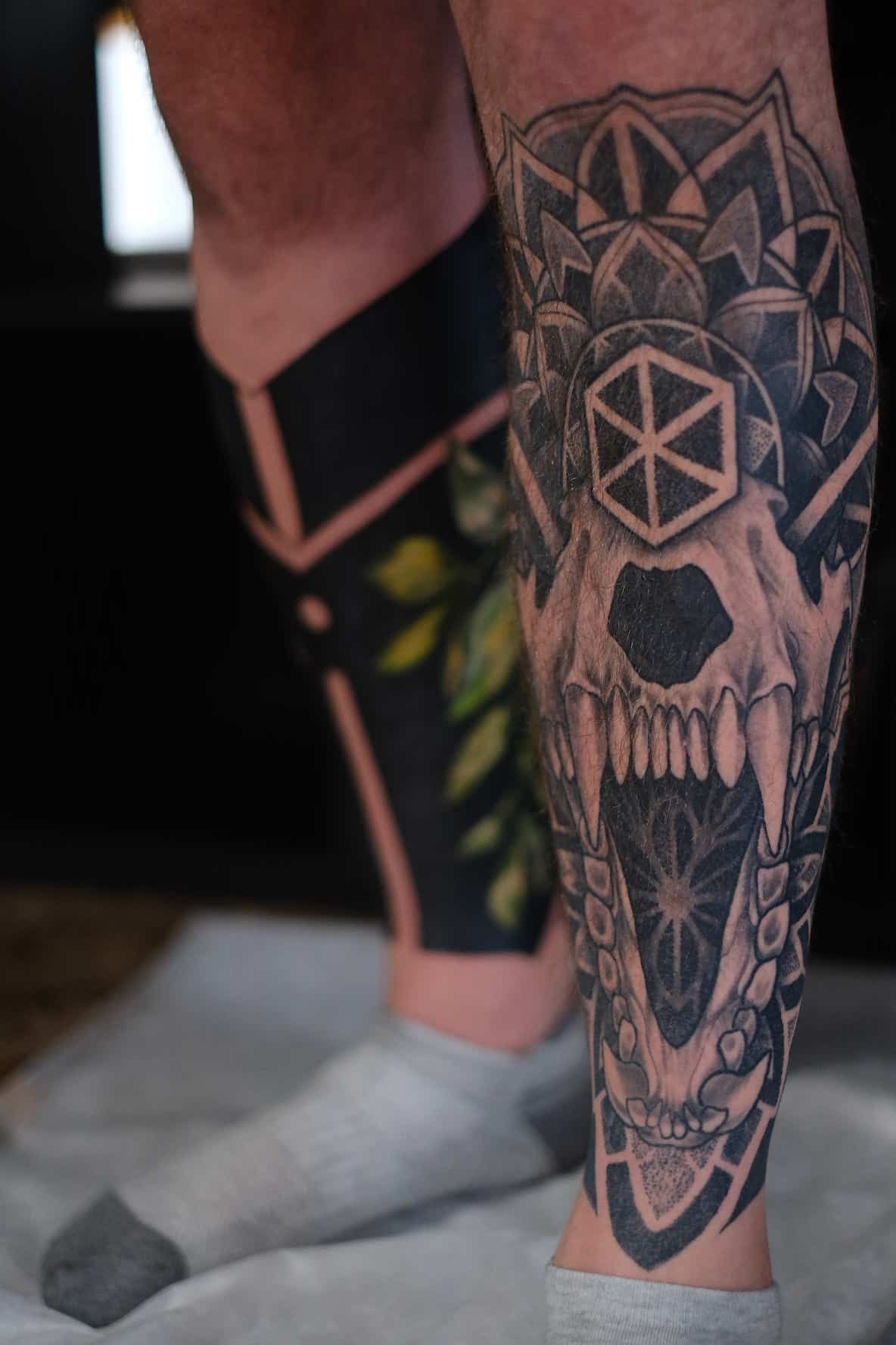 A close-up of a person's lower legs featuring intricate black ink tattoos, including a large skull design and geometry.