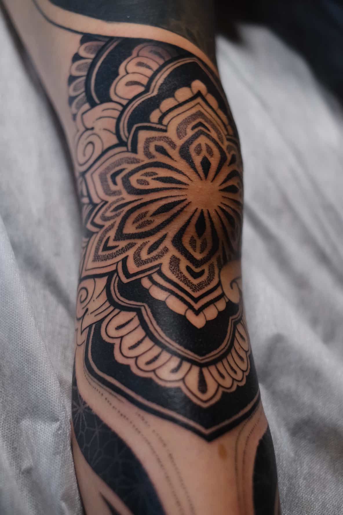 A detailed black mandala tattoo centered over a person's knee, featuring intricate geometric and floral patterns.
