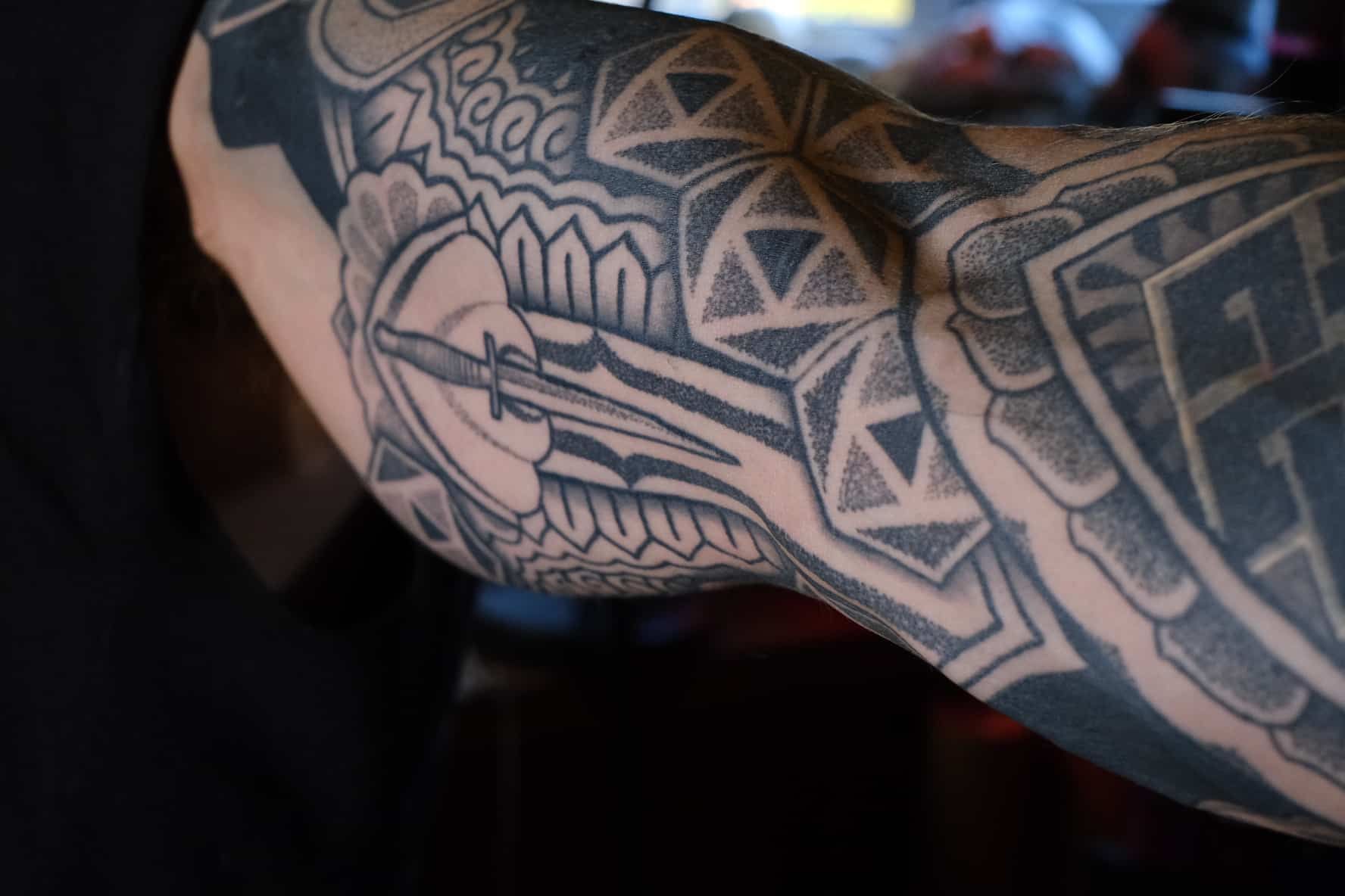 Close-up of a person's upper arm featuring detailed black-and-grey geometric patterned tattoos with a dagger.