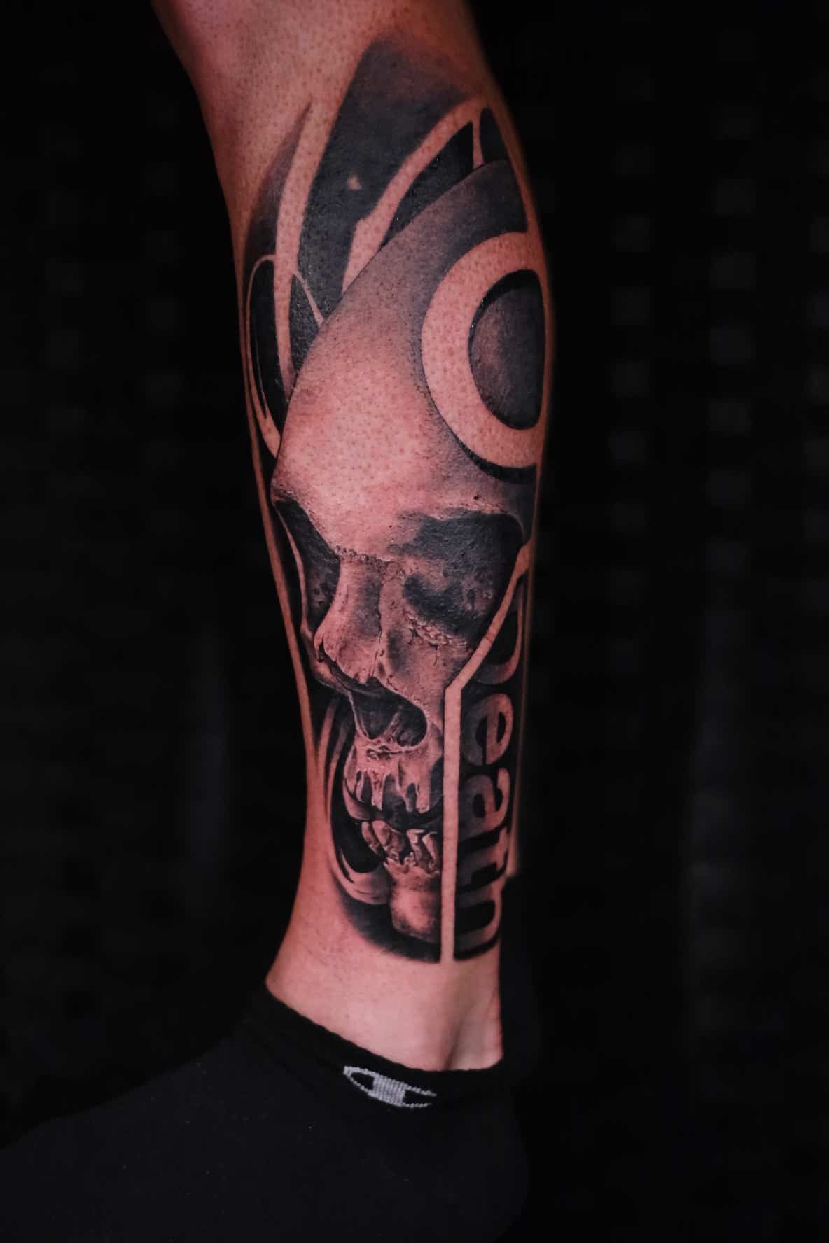 A grayscale calf tattoo of a human skull integrated into bold geometric shapes and the word 