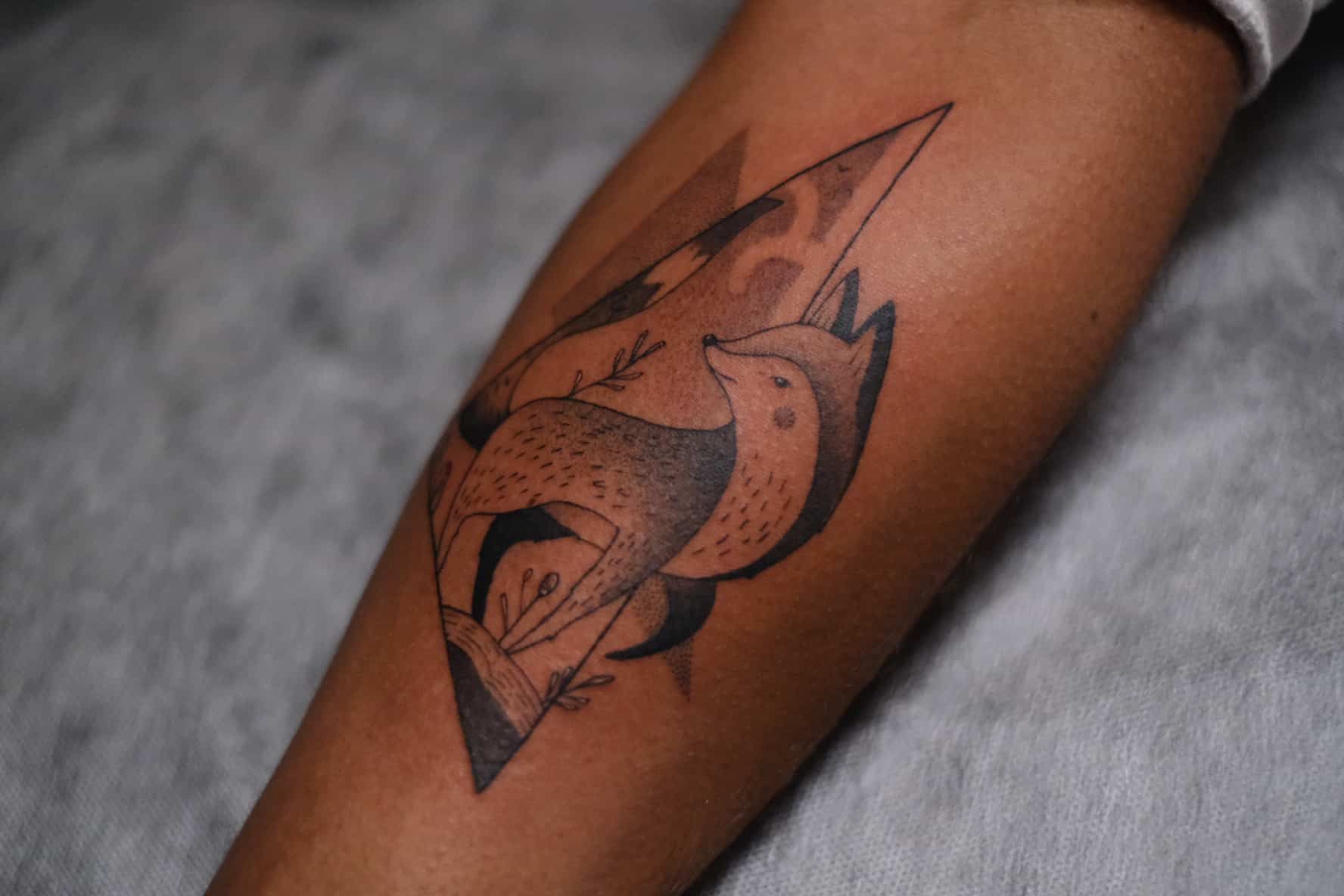 A black ink tattoo on an arm featuring a stylized fox enclosed within a diamond-shaped geometric border.