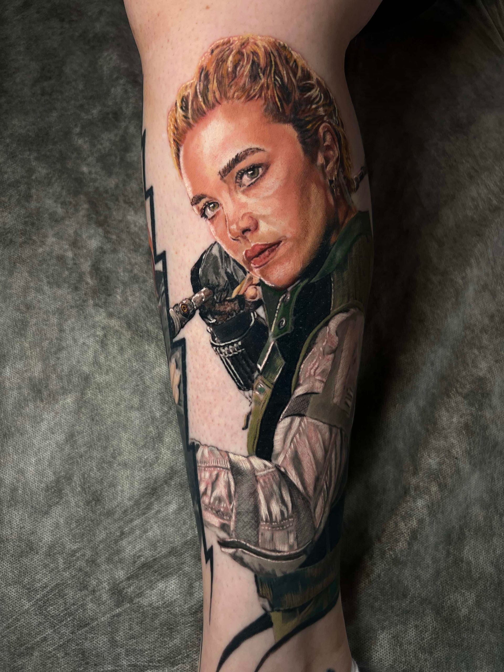A realistic color tattoo of Florence Pugh as Yelena Belova on a person’s lower leg.