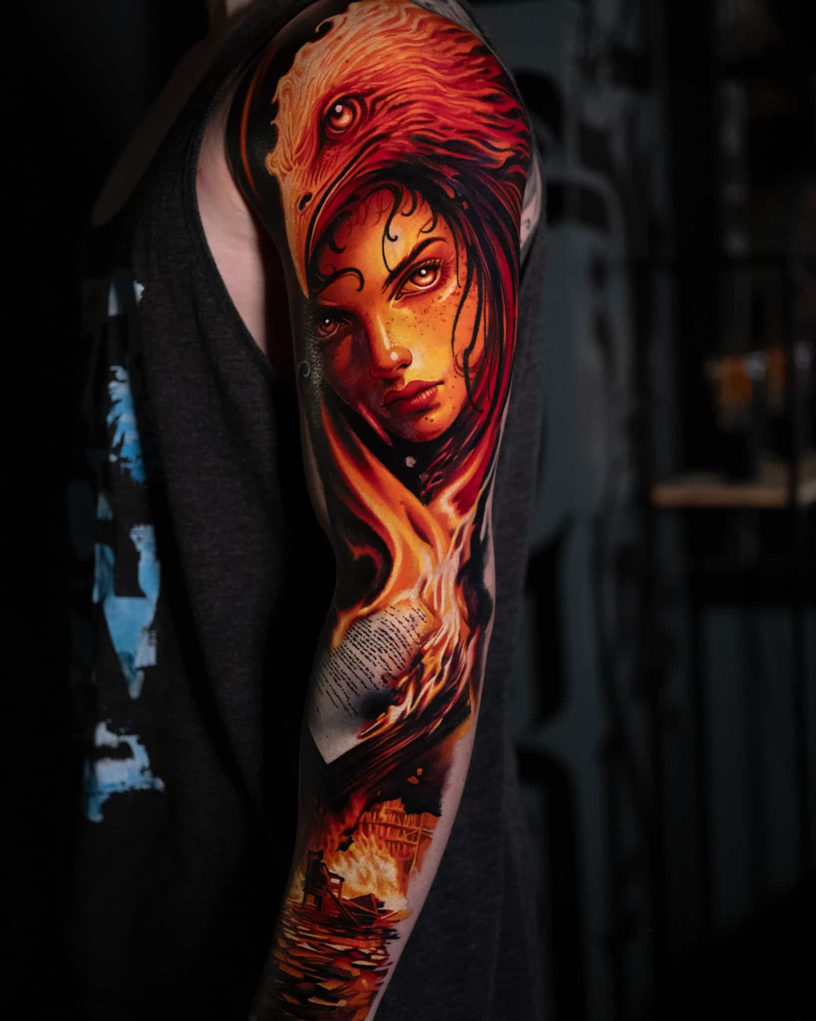 A vibrant, detailed tattoo of a phoenix head merging into a face on an arm, featuring warm orange, red, and dark tones.