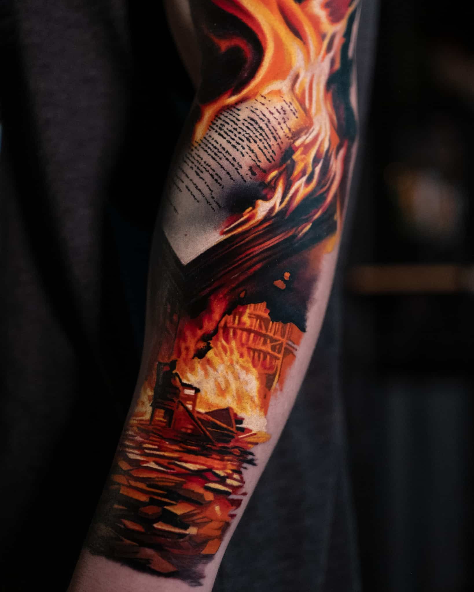 A colorful arm tattoo depicting a burning document floating above a landscape engulfed in bright, orange-red flames.