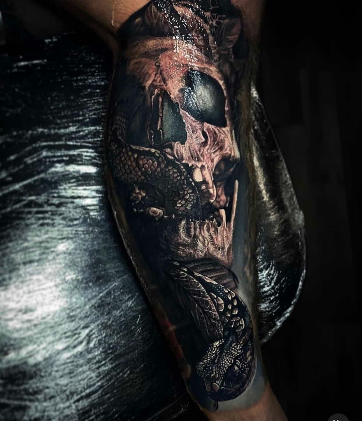 Detailed dark-themed tattoo on an arm, featuring a realistic human skull intertwined with a coiled snake.