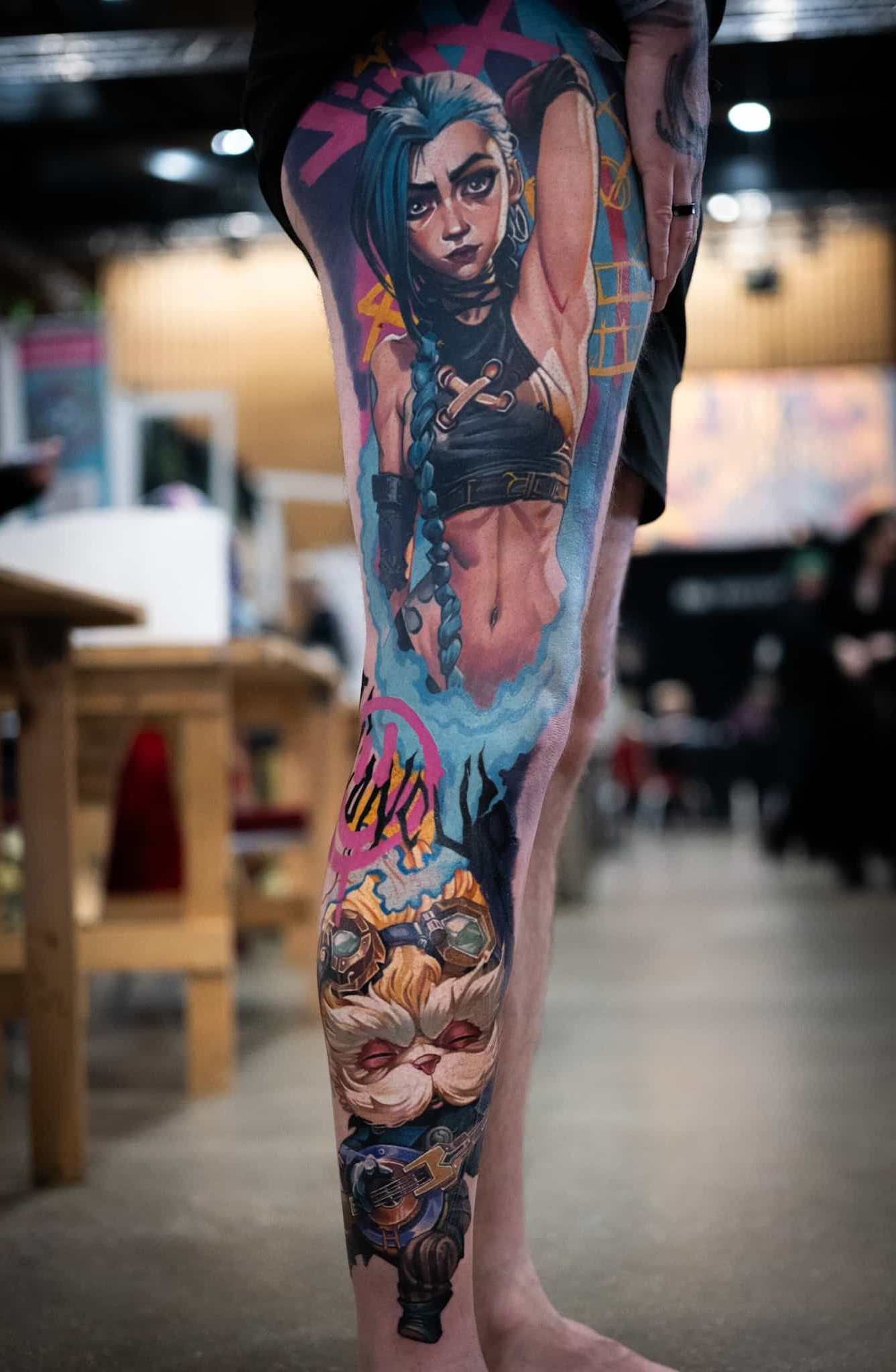 A leg tattoo featuring Jinx from Arcane, with blue hair and stylized artwork, standing in a blurred room.