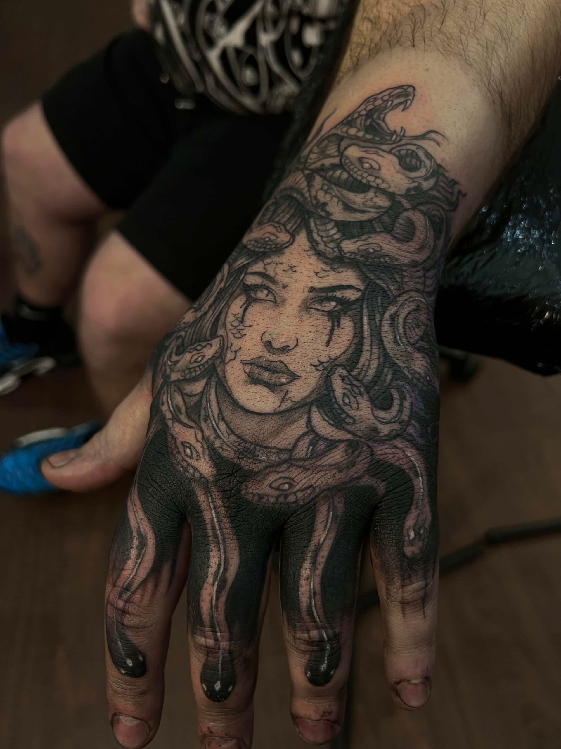 A detailed grayscale tattoo of Medusa covering the back of a hand, featuring coiled snakes and dark, stippled shading.