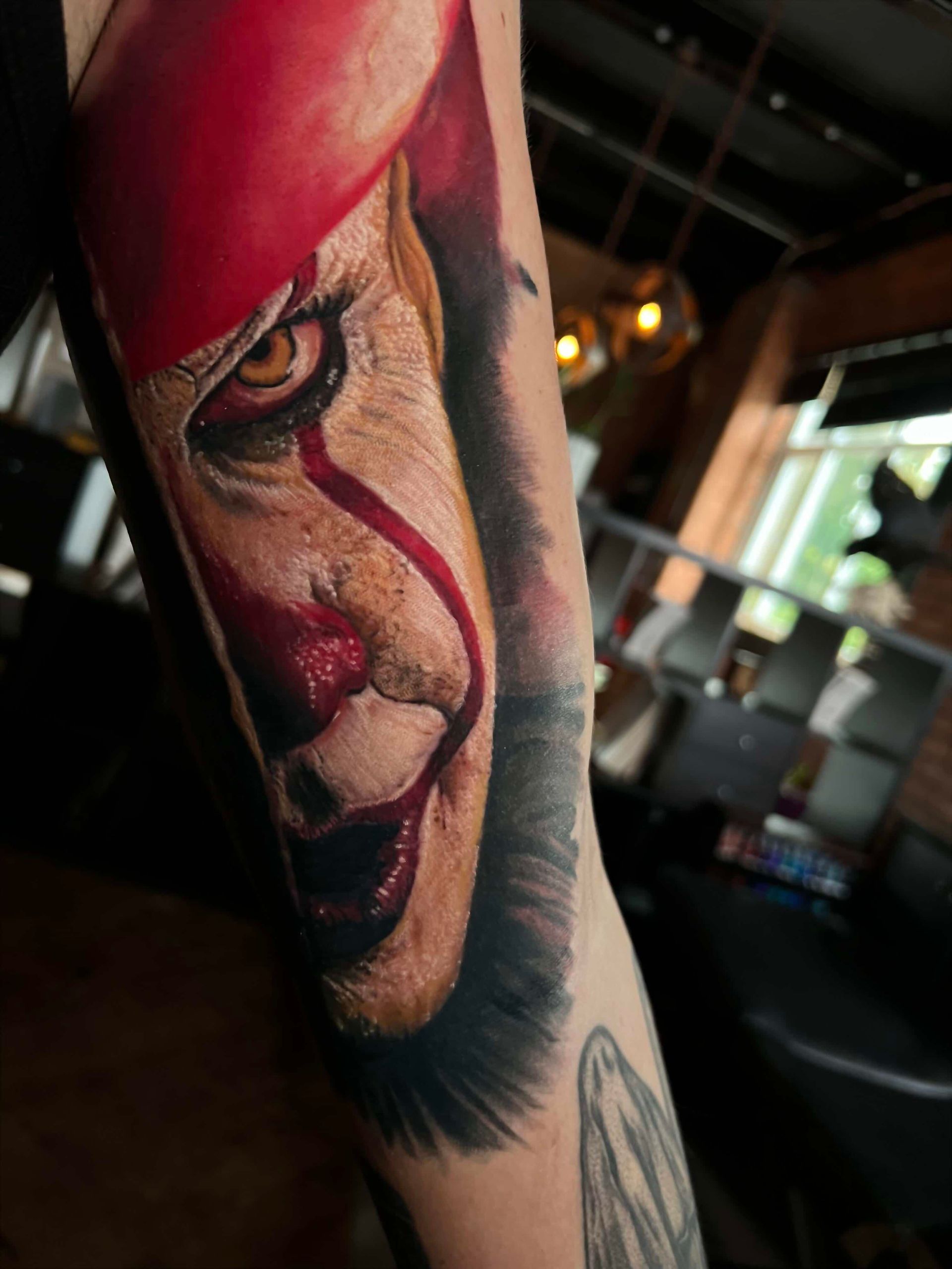 A detailed, realistic tattoo of Pennywise the Clown featuring bright red makeup and intense, golden-colored eyes.